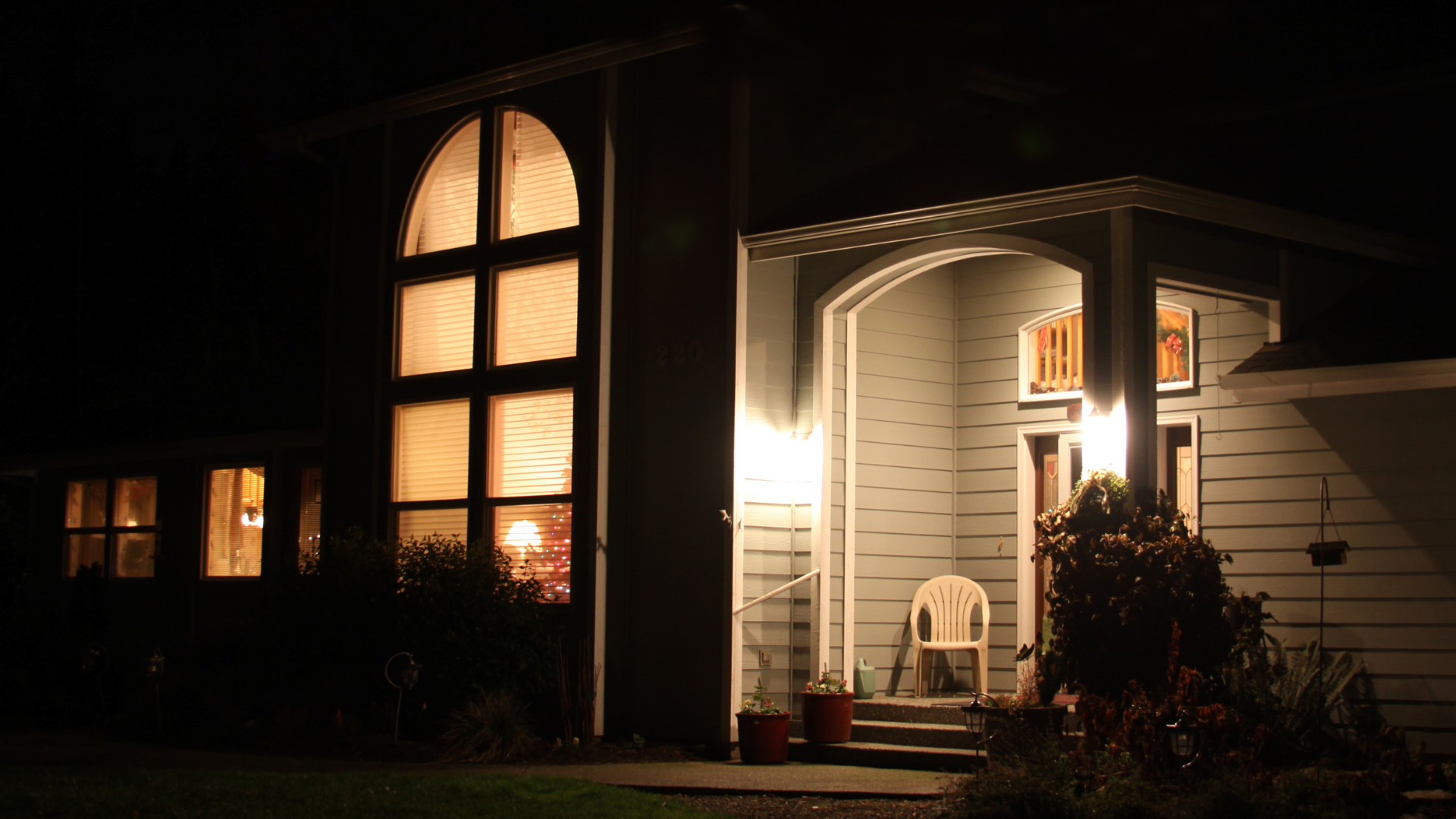 Night view of a house. Illuminated windows and porch with a white chair, set against a dark sky.