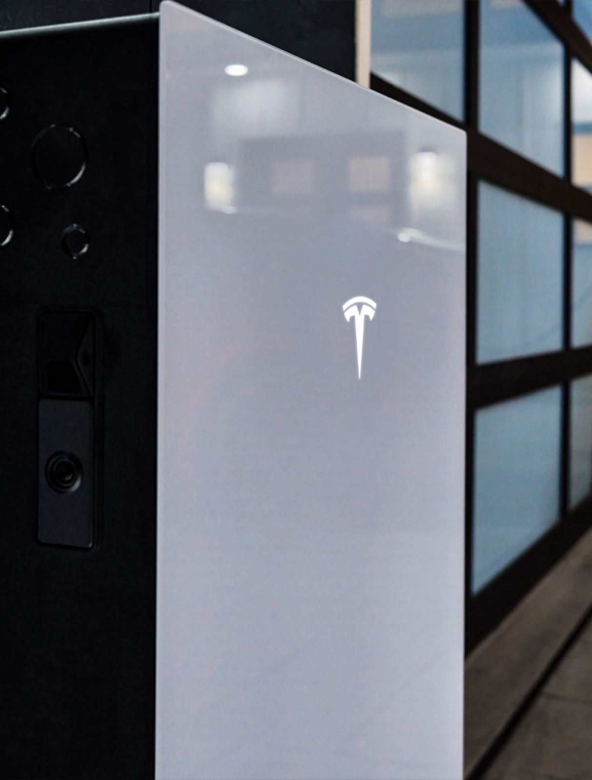 Tesla Powerwall unit, gray front with logo, next to black electrical panel, garage background.