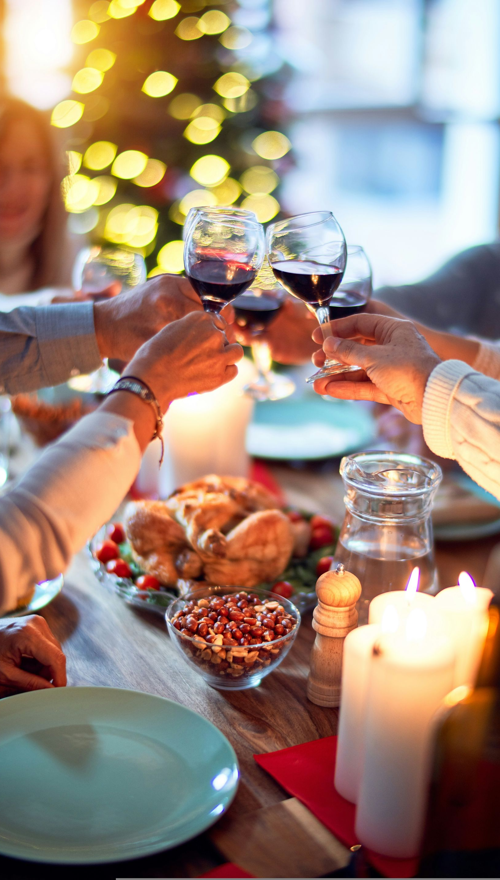 People toasting with wine glasses at a Christmas dinner table, lit by candles and a decorated tree.