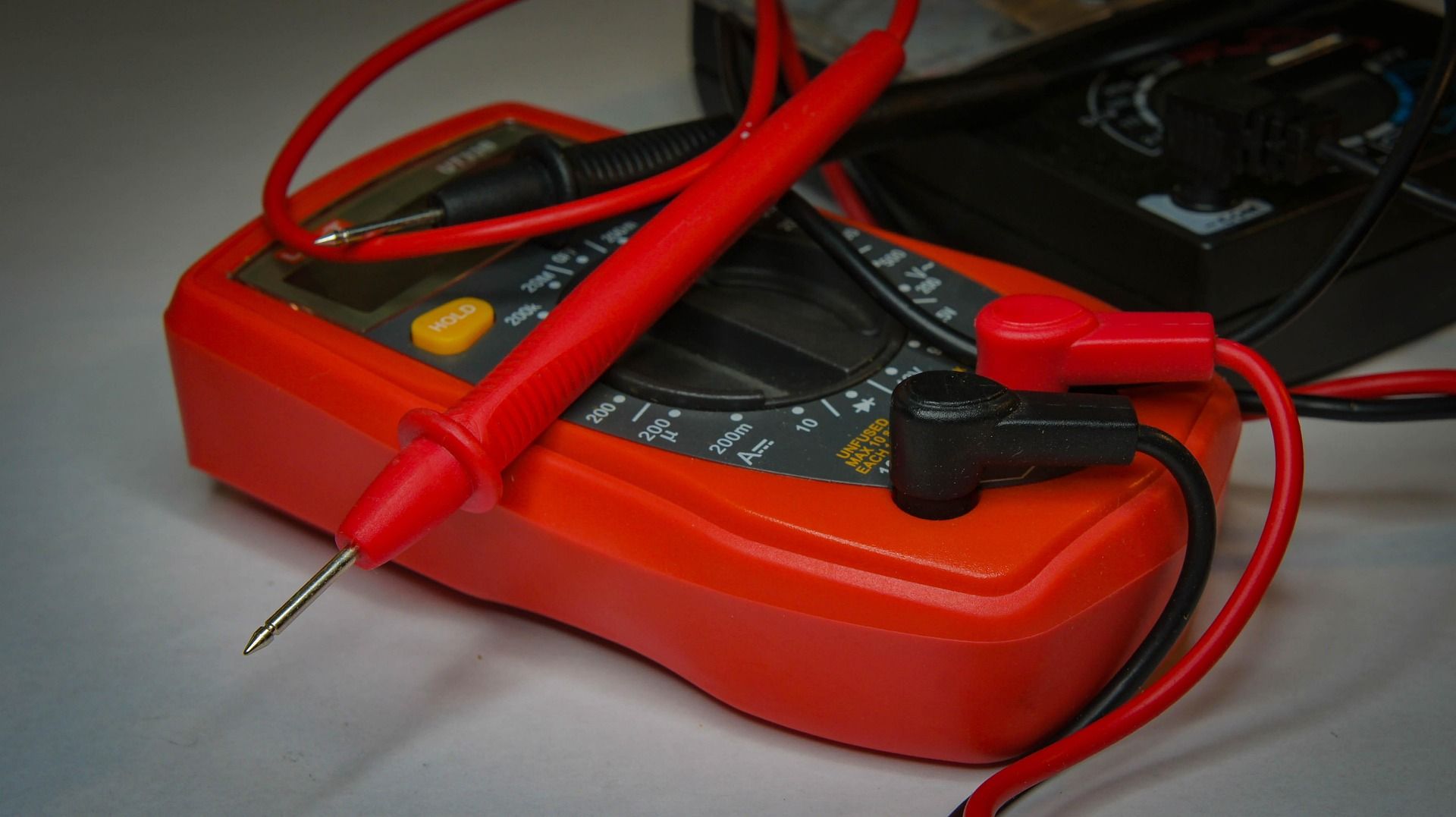 Red multimeter with probes, black knobs, and digital display, on a light surface.