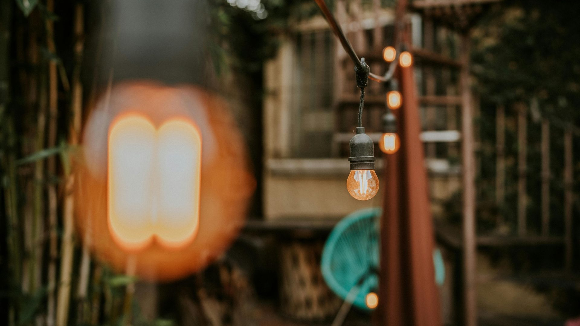 String of illuminated Edison-style bulbs hanging outside; background includes a spiral staircase and greenery.