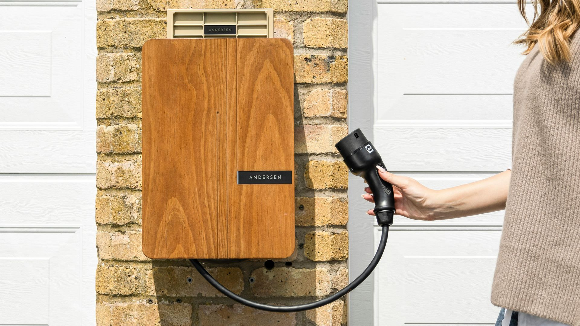 Woman holding EV charger, plugged into a wooden charging station on a brick wall. White garage door in background.