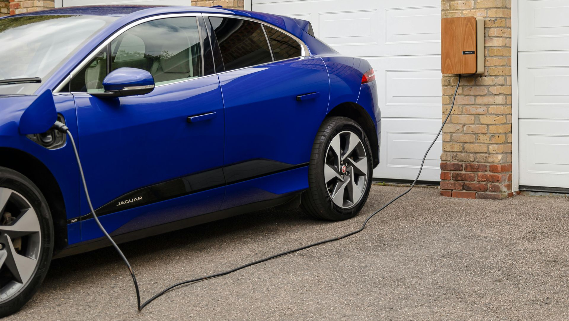 Blue electric car charging at a home garage with a charger mounted on the brick wall.