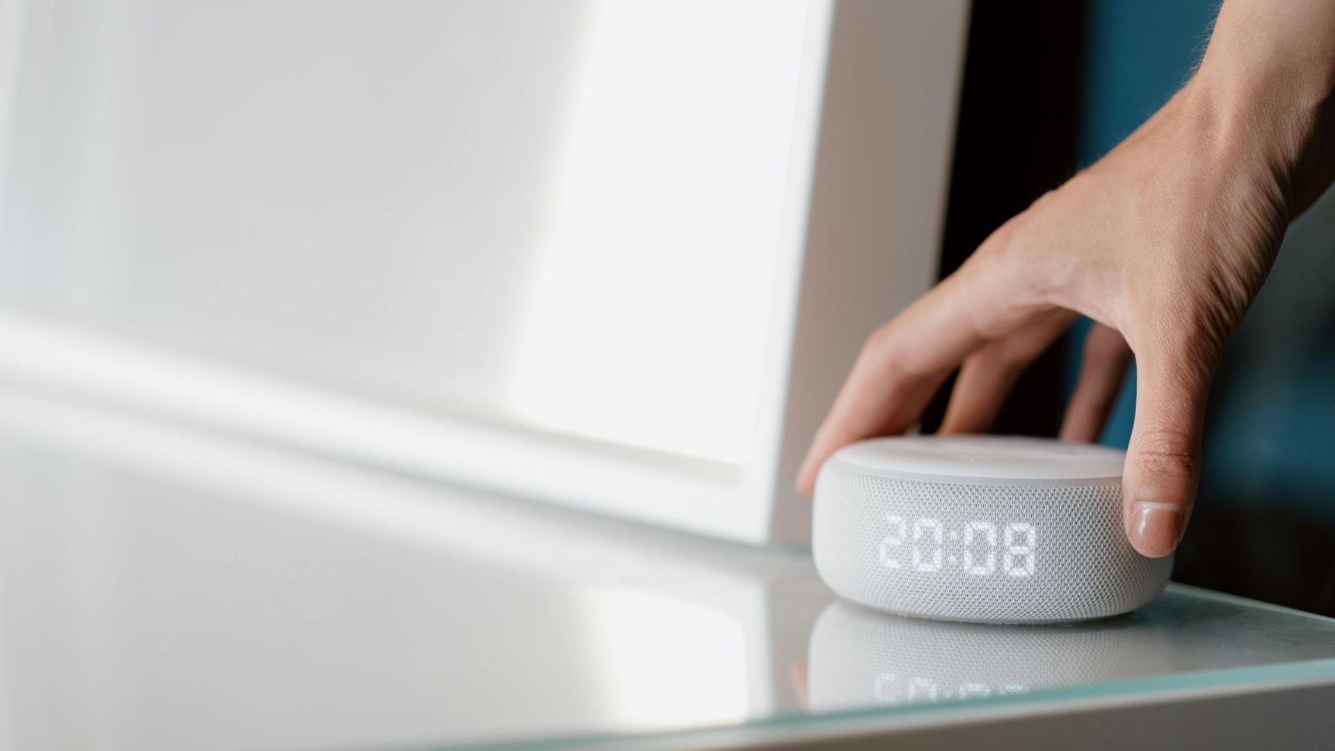 Hand reaching for a small, circular digital clock displaying 20:08 on a glass table.