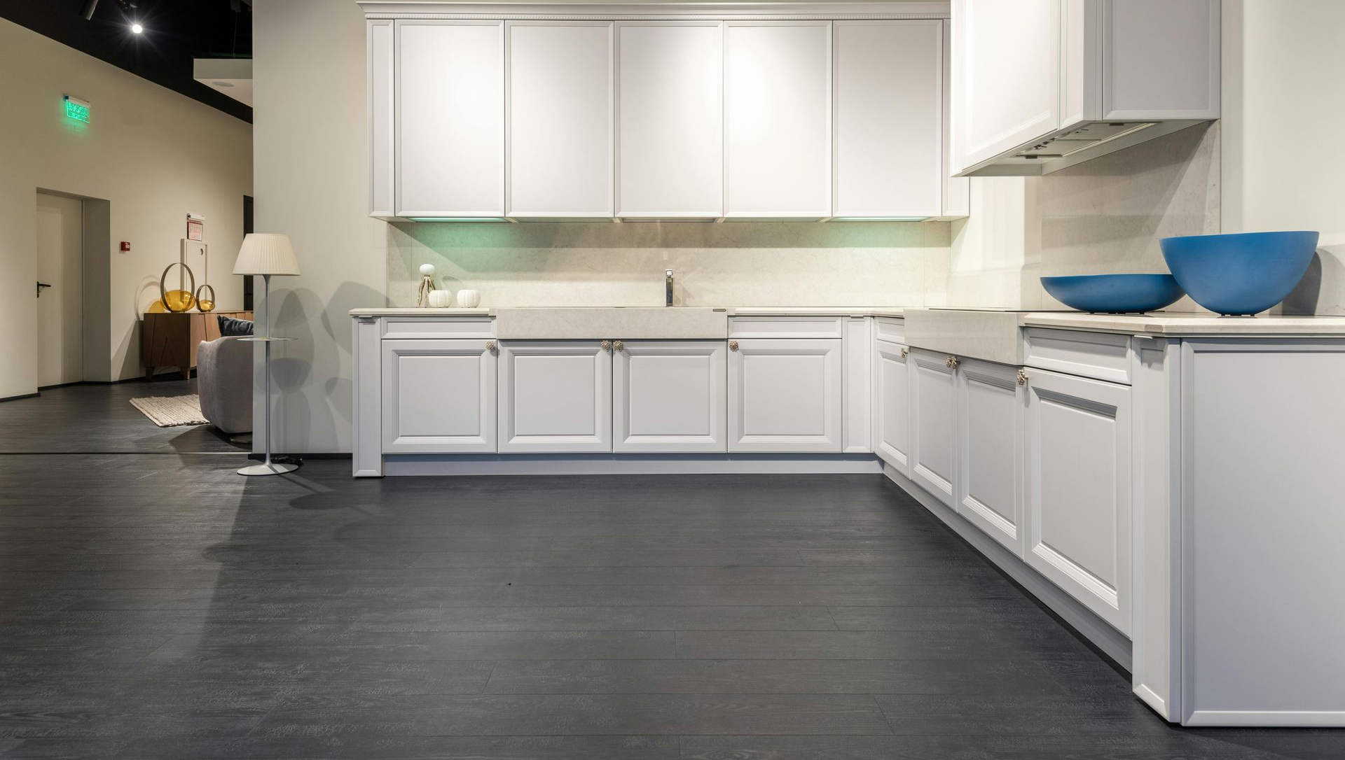 A white modern kitchen showroom with white cabinets, light countertops, a farmhouse sink, and dark wooden flooring.