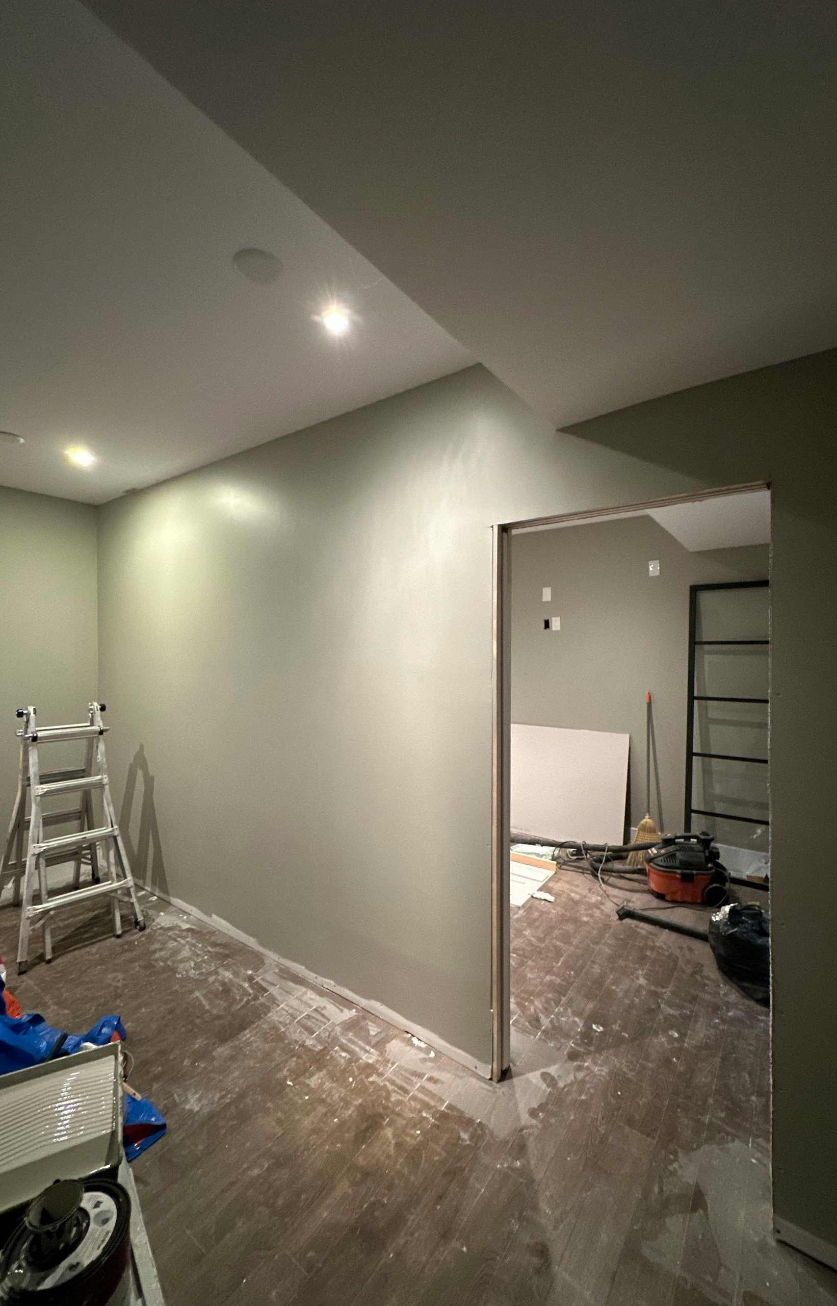 A room under renovation with sage green walls, recessed lighting, a stepladder, and an open doorway to another space.