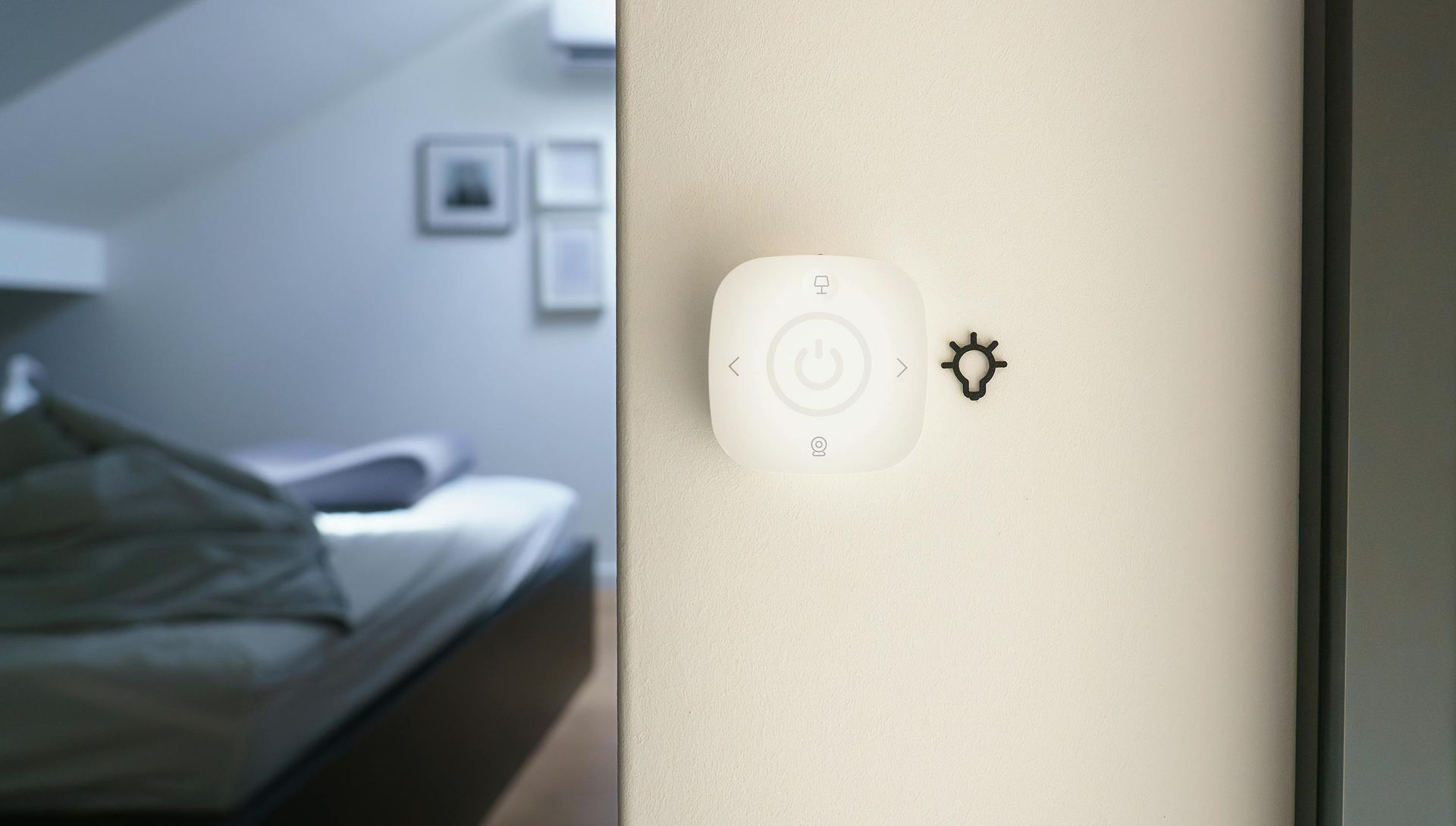 A white wireless smart light switch mounted on a light-colored wall, with a blurred bedroom interior in the background.