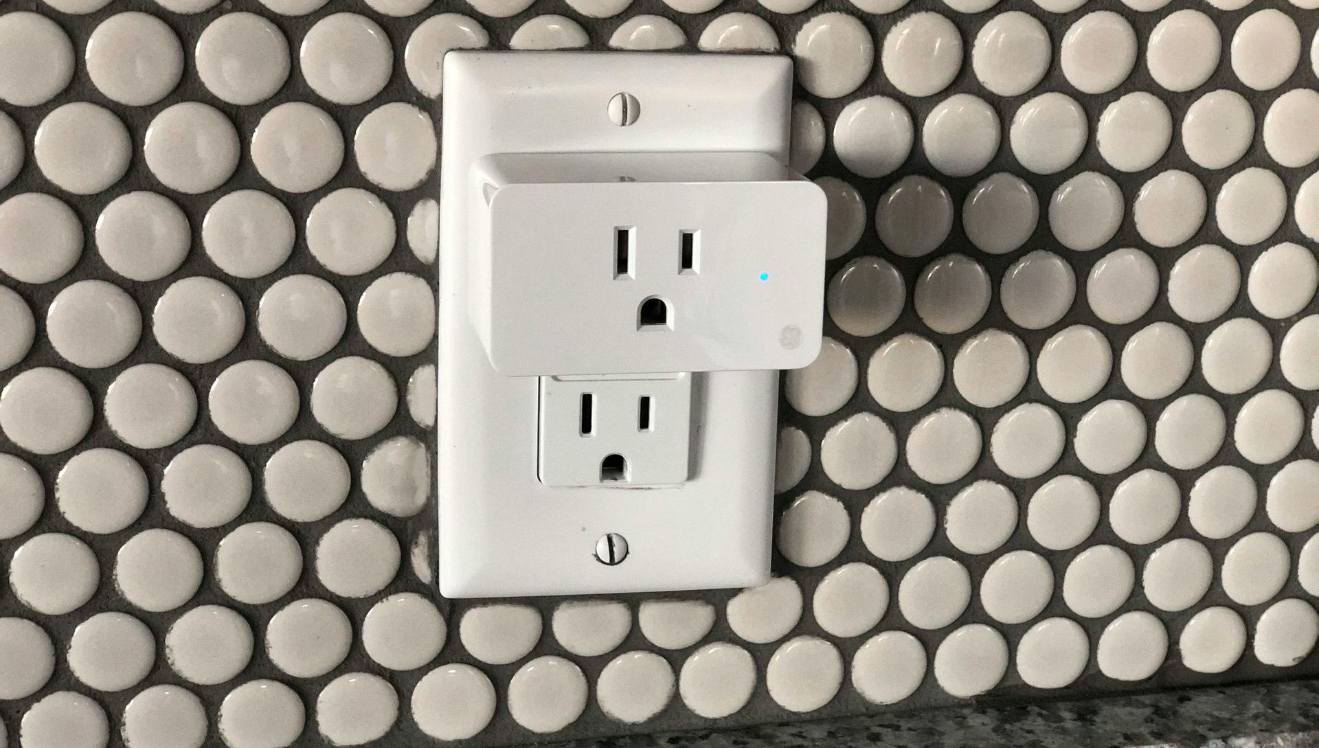 A white smart plug adapter is plugged into the top outlet of a wall receptacle against a white penny tile backsplash.