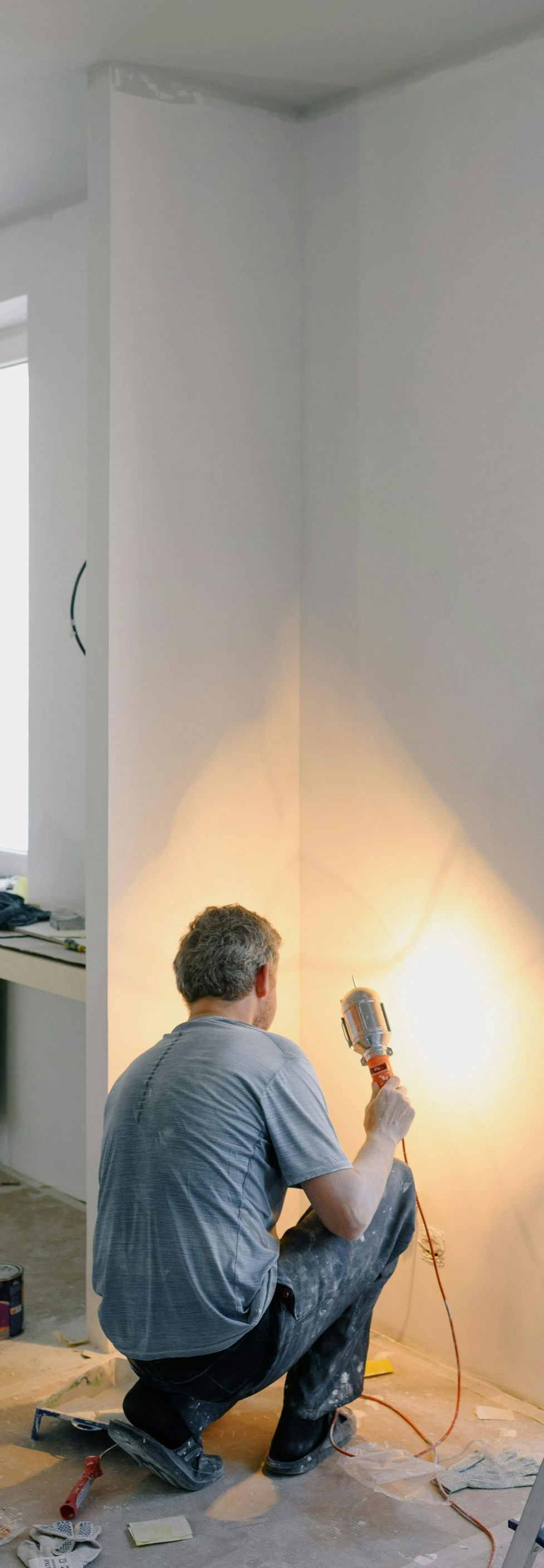 A person in work clothes kneels while using a handheld light to inspect a freshly painted white wall in a room.