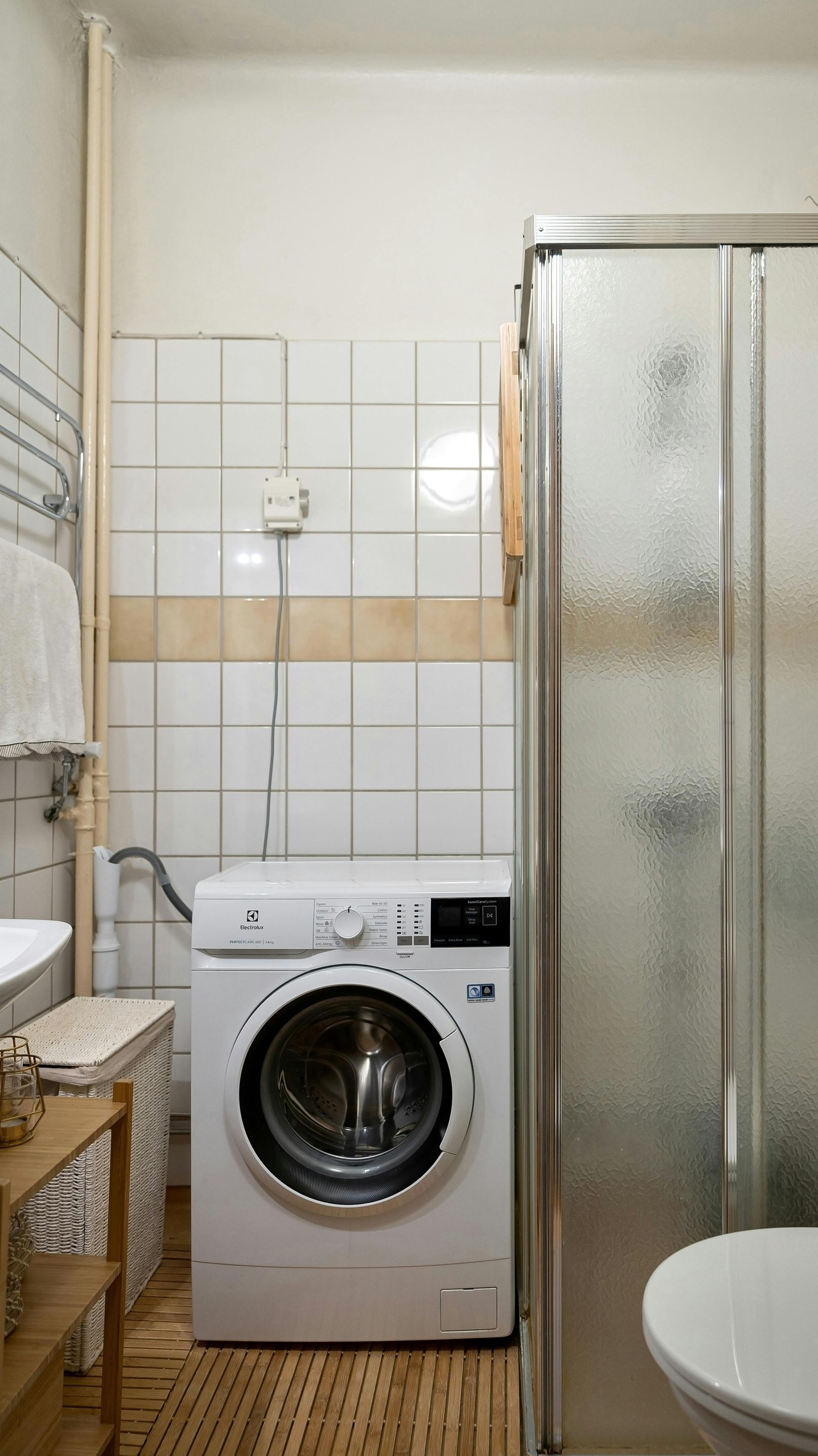 Small bathroom with washing machine, shower stall, and toilet. White and beige tiles, wooden floor.