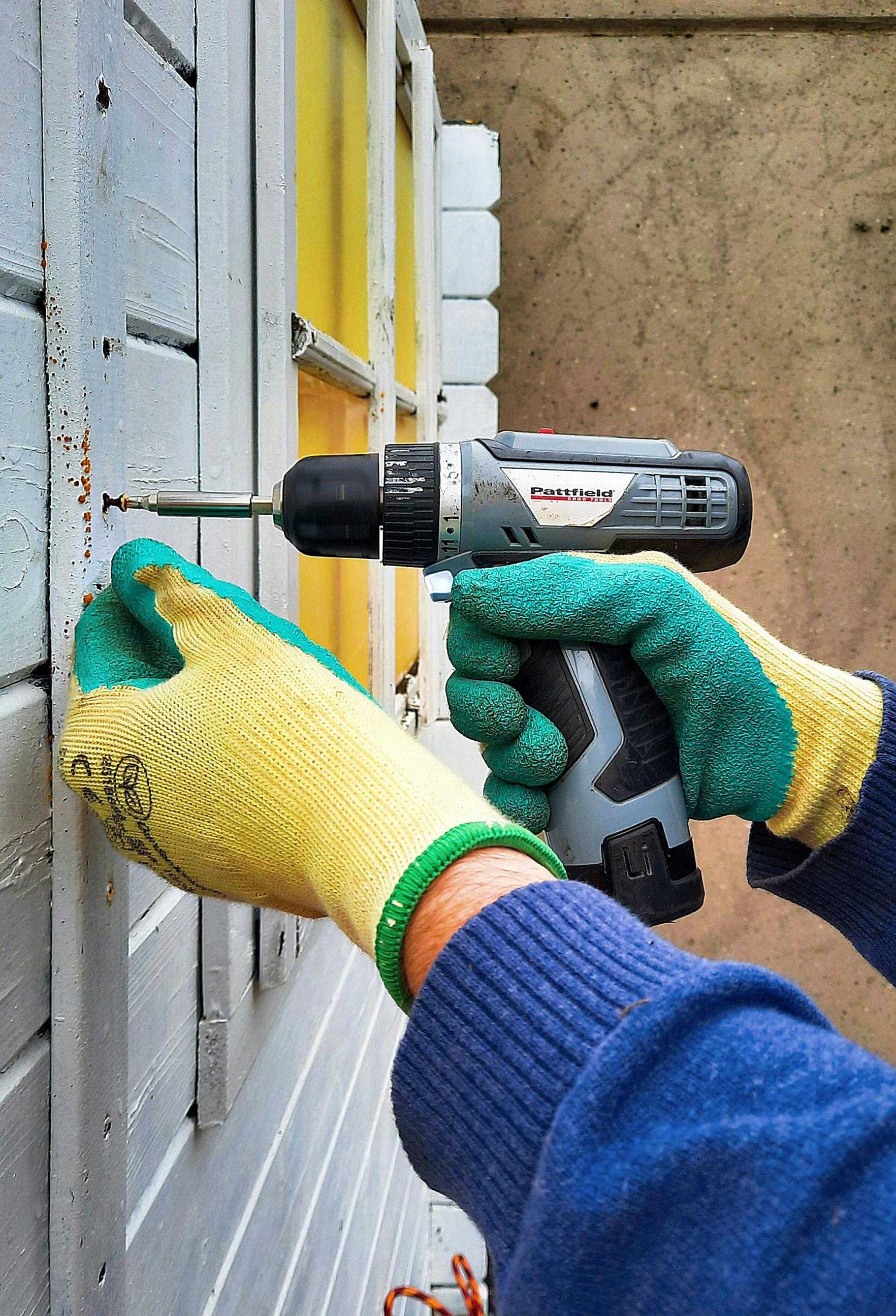 Hands wearing green and yellow work gloves use a power drill to drive a screw into a gray wooden wall.
