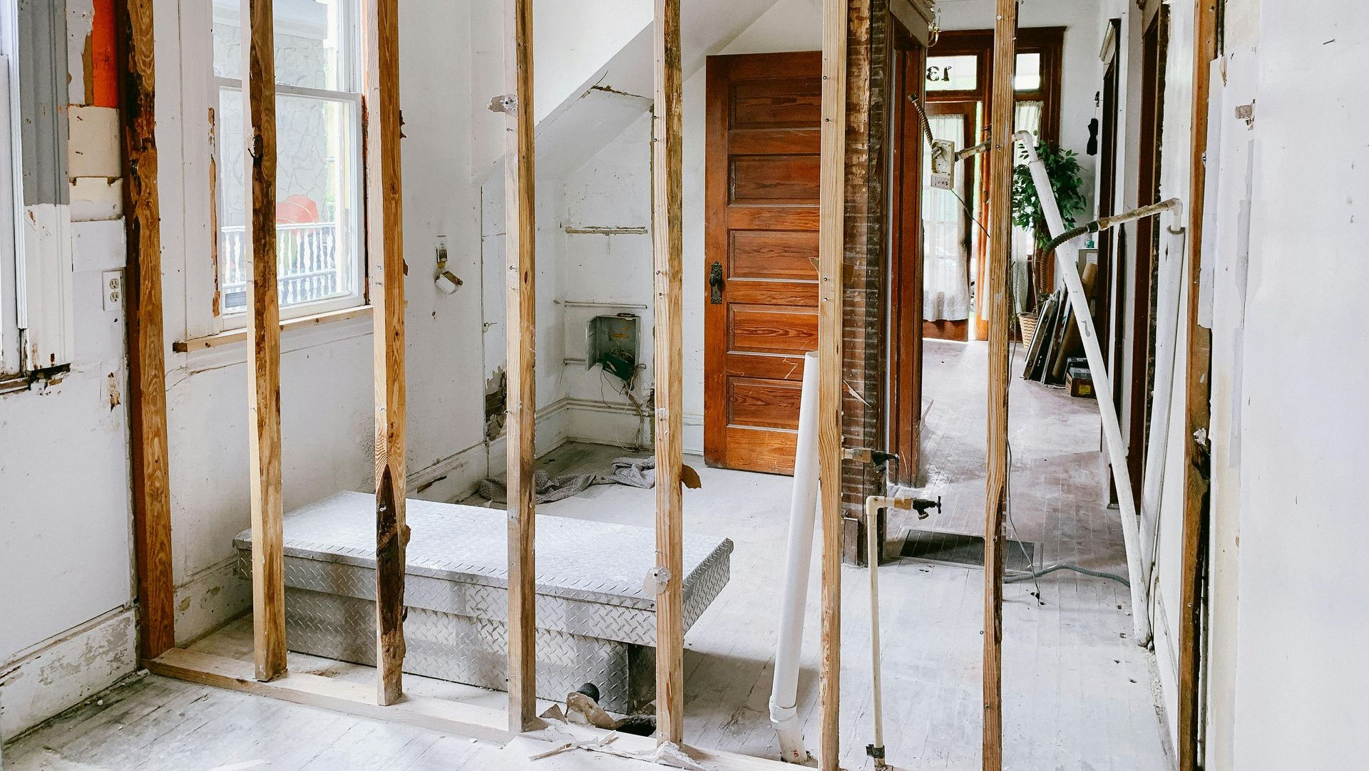 Interior view of a room under renovation with exposed wooden wall studs, white plaster walls, and a wooden door.