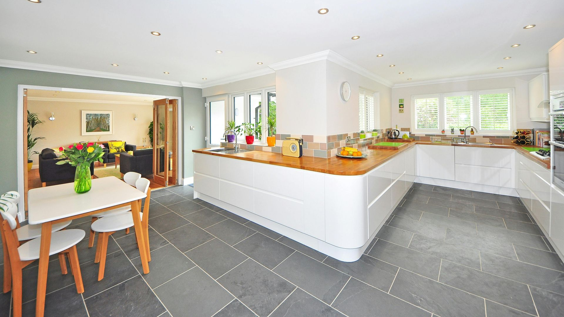 Bright, open-plan kitchen with white cabinets, wooden countertops, and gray tile flooring. Dining table in foreground, living room visible.