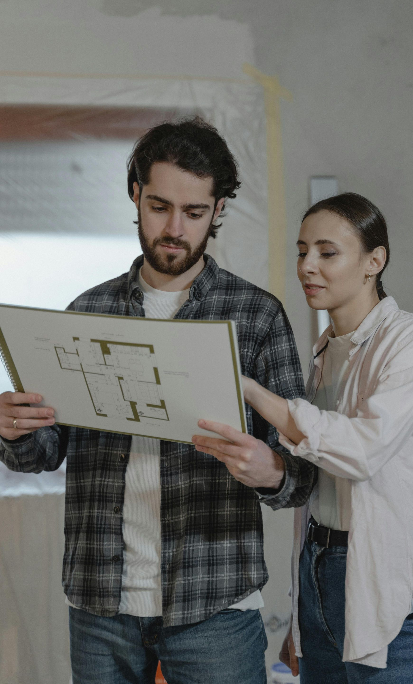 A man and woman in a room under construction hold and review a printed floor plan together.