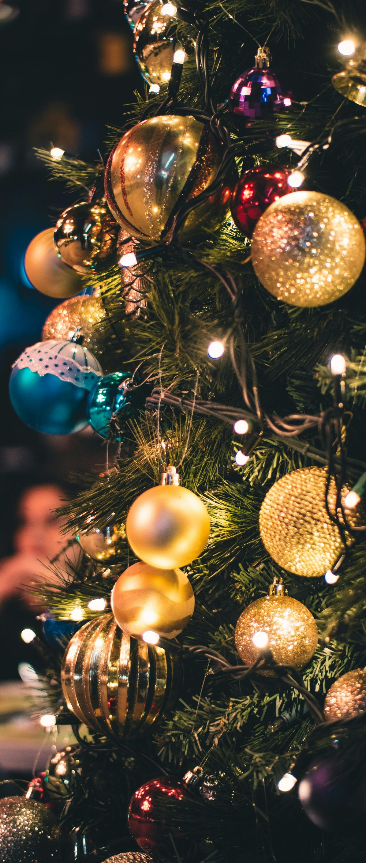 Christmas tree decorated with gold, teal, and red ornaments, illuminated by white lights.