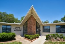 A clean, bright medical consultation office with two diagnostic machines, white desks, rolling chairs, and large windows.