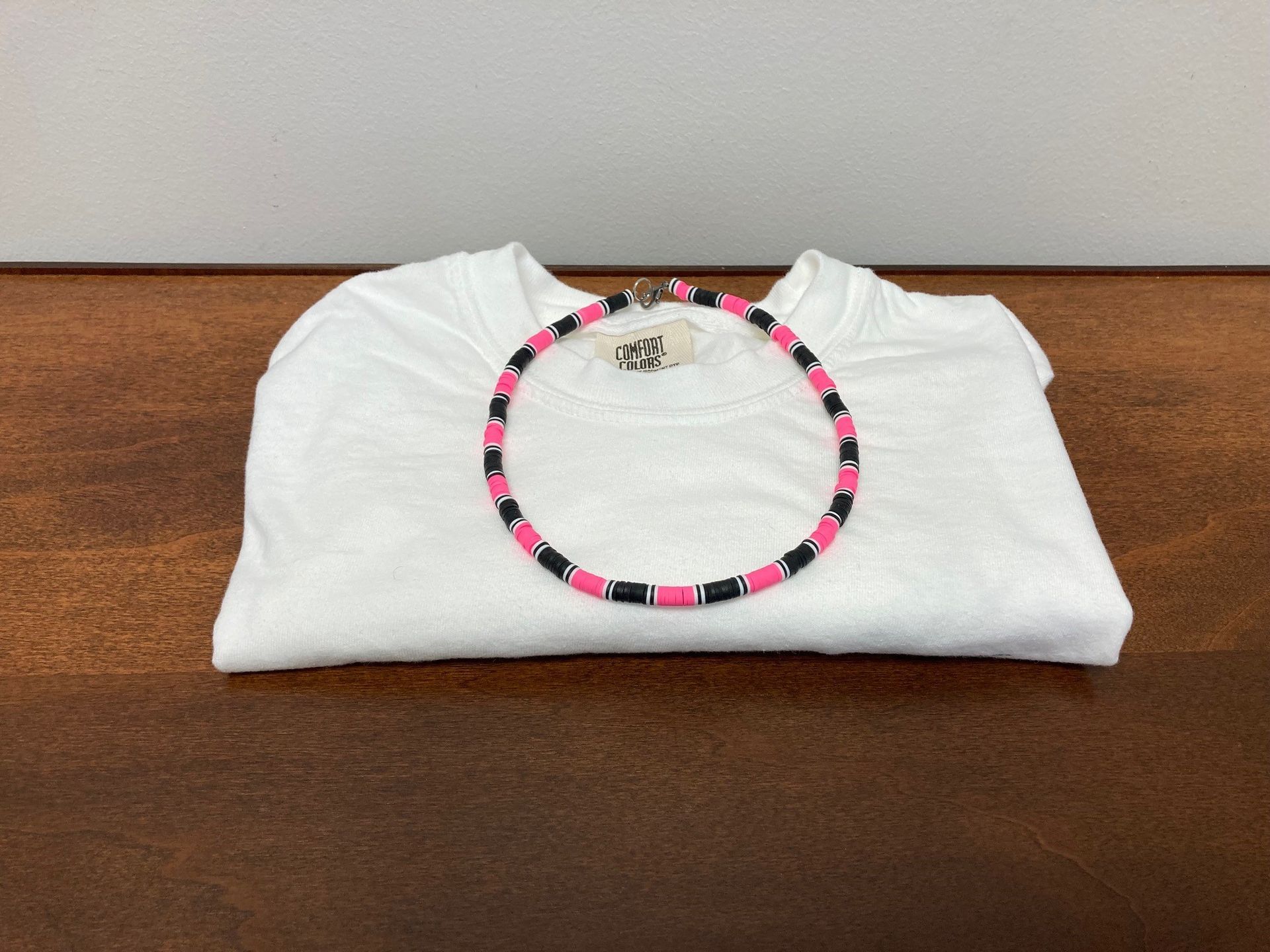 Folded white T-shirt on a wooden table with a black-and-pink beaded necklace on top