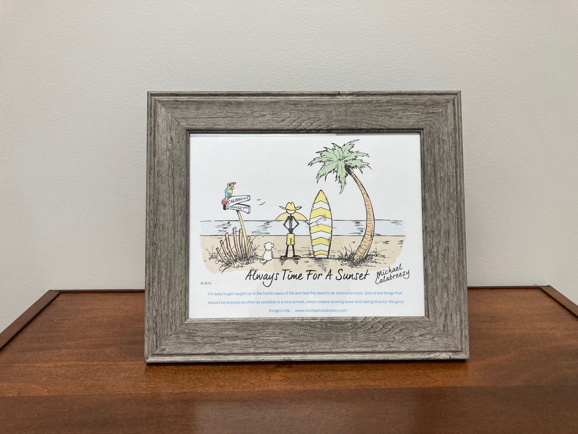Framed beach-themed artwork on a wooden table with beach chairs, palm tree, and surfboards