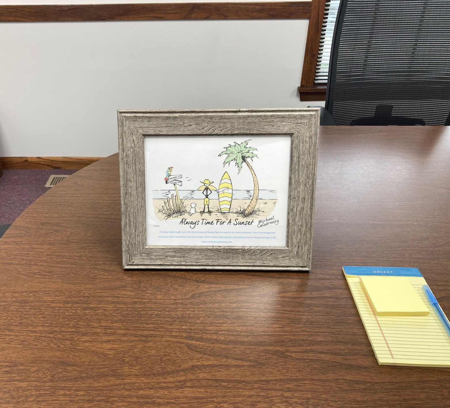Framed beach-themed artwork on a wooden desk beside a yellow sticky-note pad.
