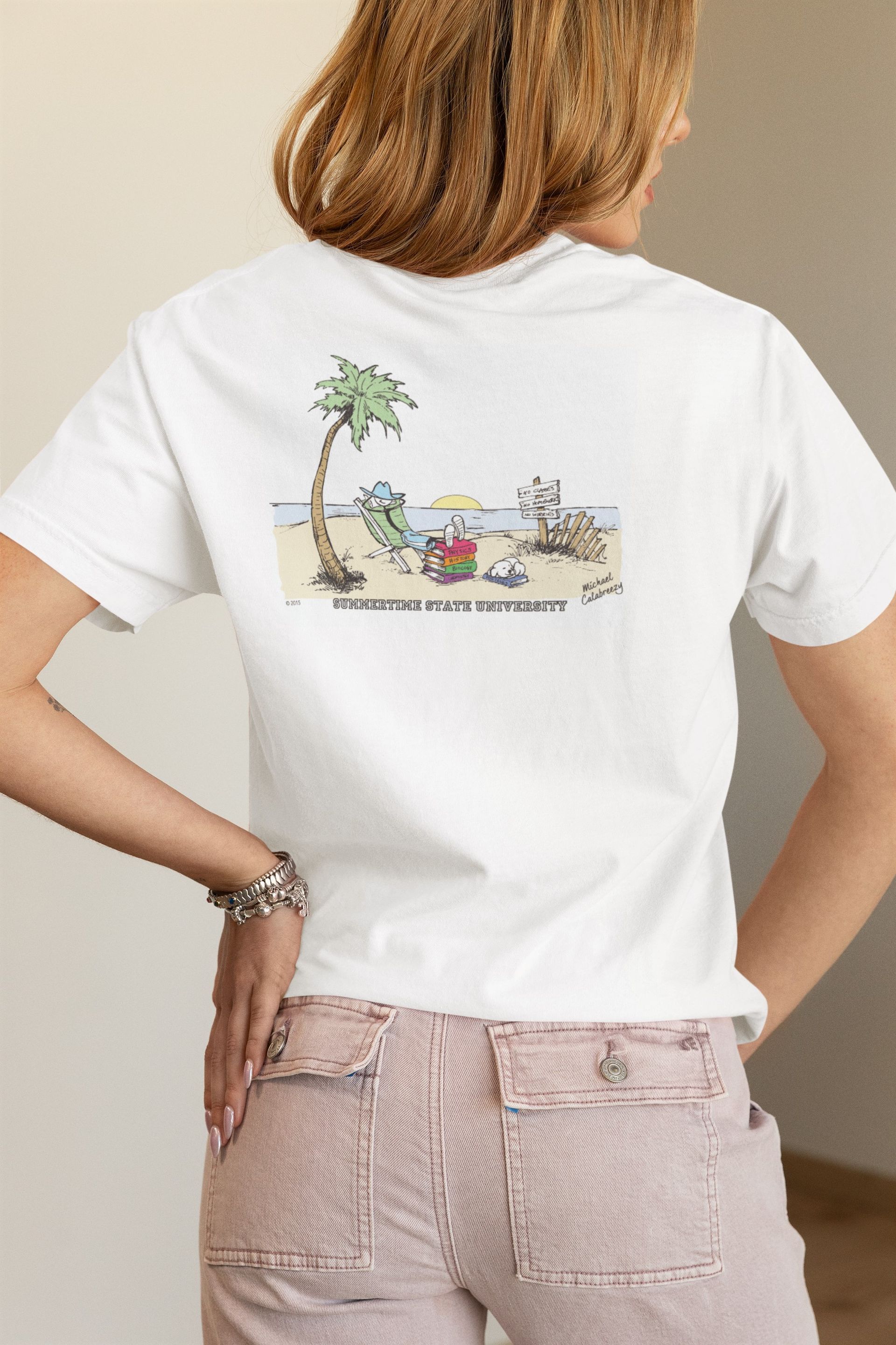 Person in a white T-shirt with a small beach scene graphic on the back and pink striped pants, standing indoors.