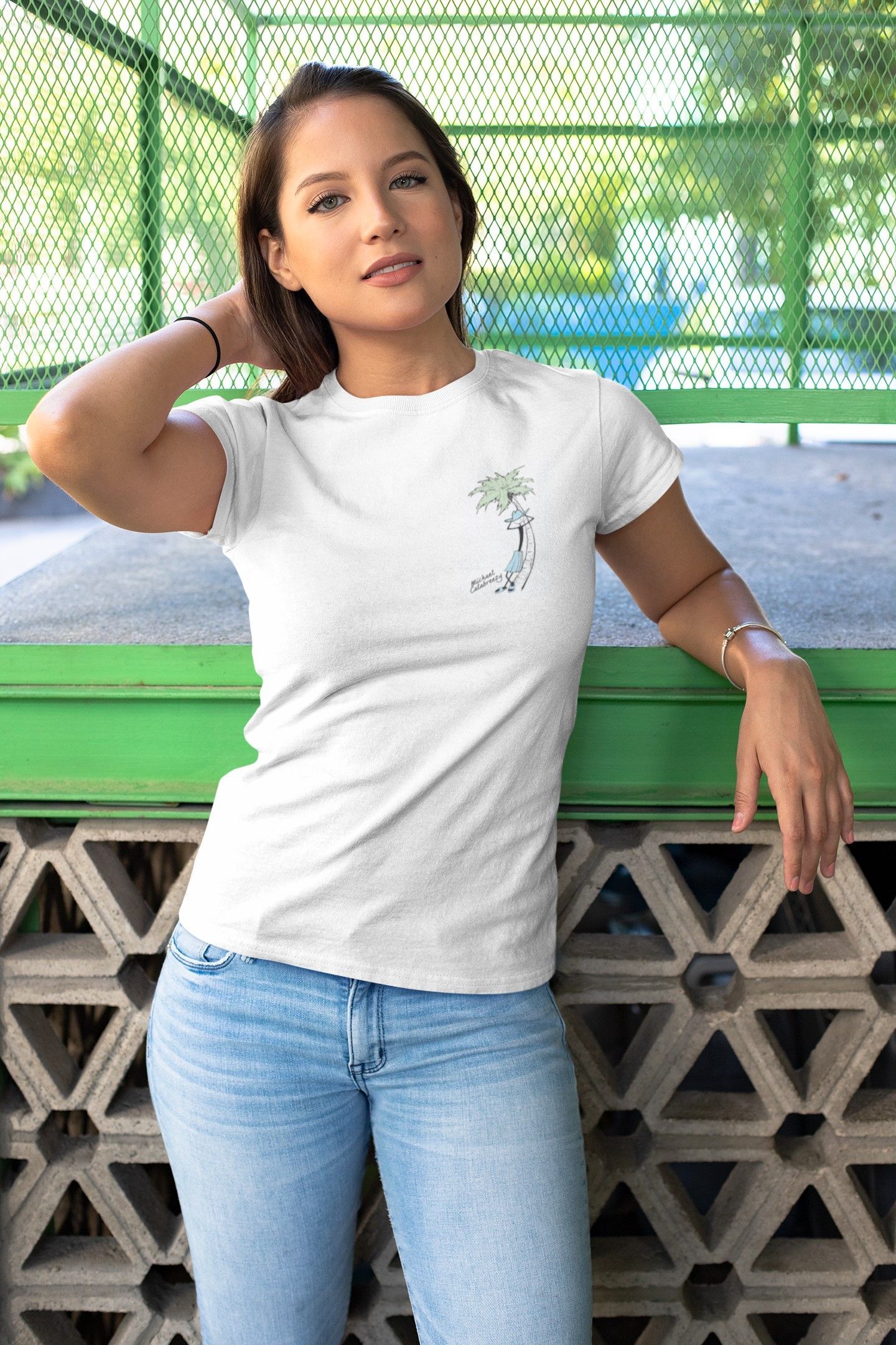 Woman in a white T-shirt and jeans posing outdoors against a green bench and patterned wall.