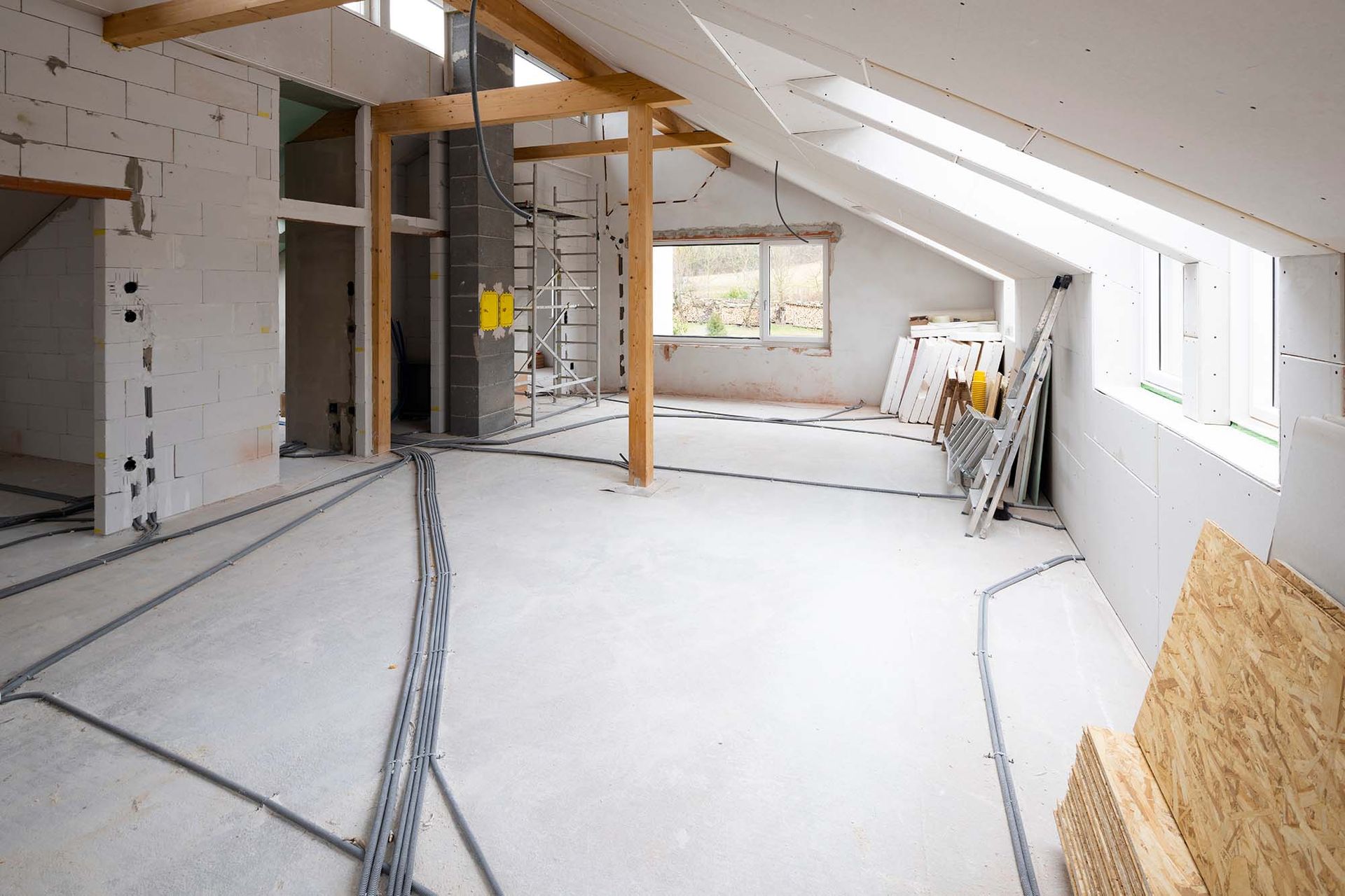 Unfinished attic room with white walls, exposed beams, windows, and construction materials on the floor.