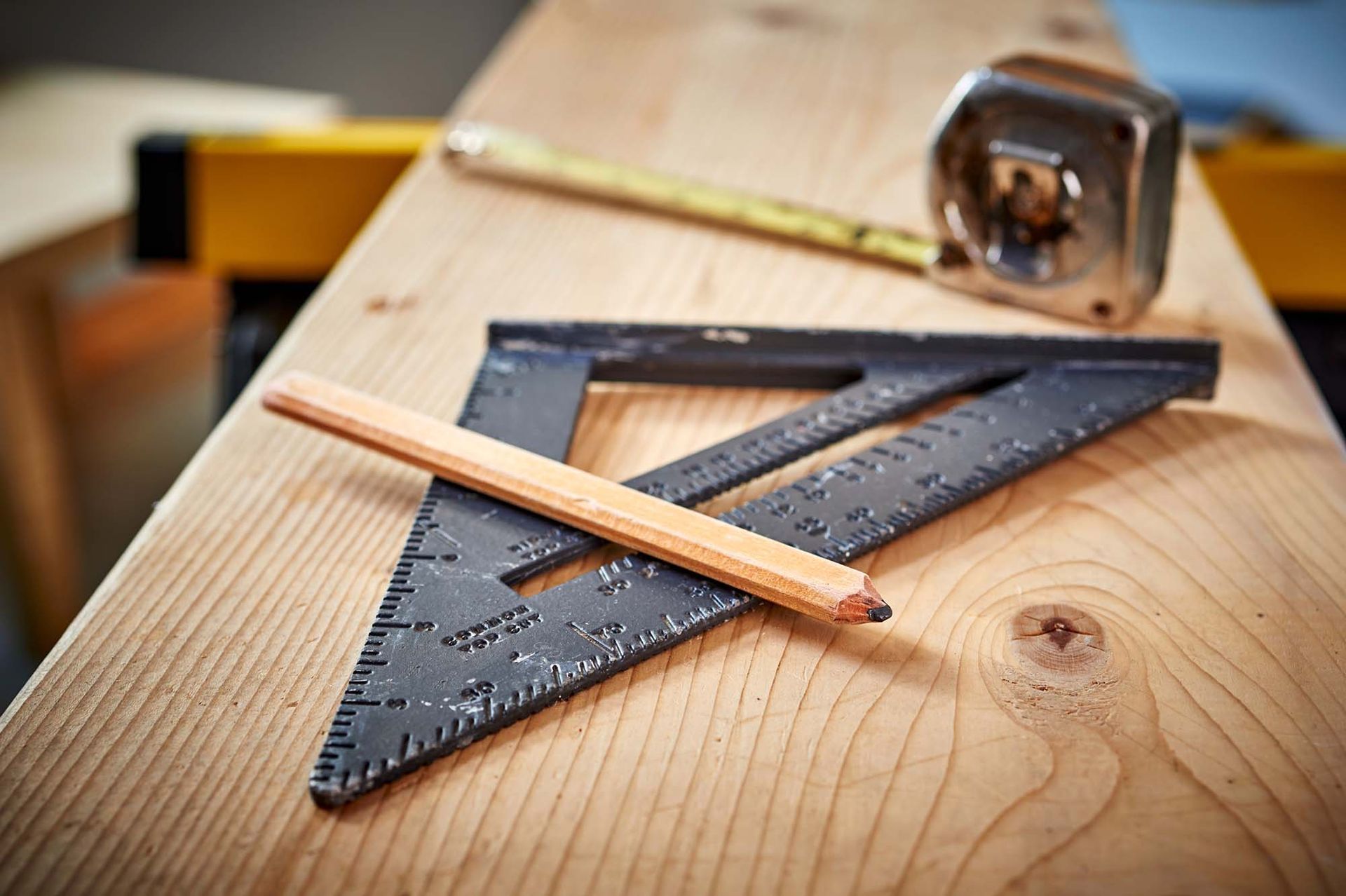Pencil on a black drafting triangle and ruler on a wooden workbench with a tape measure in the background