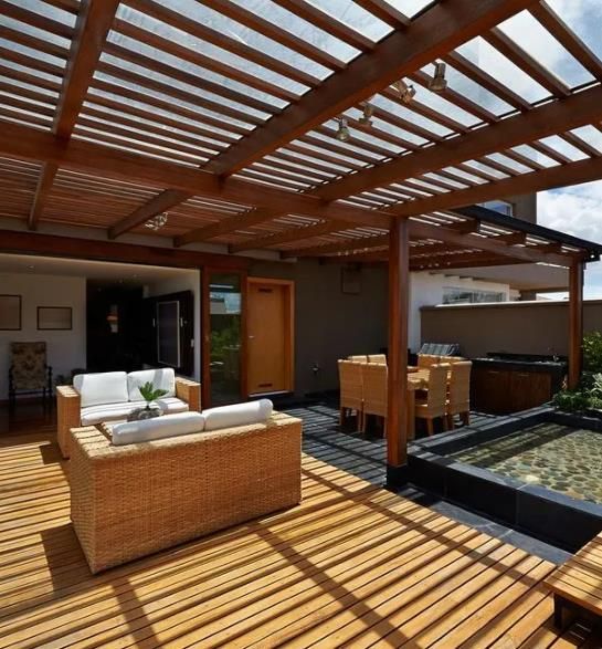 Covered wooden patio with wicker seating, dining table, and sunlit pergola shadows overlooking a yard