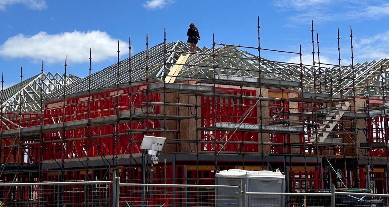 Scaffolding around a building under construction, with red safety netting and a partly roofed frame under a blue sky