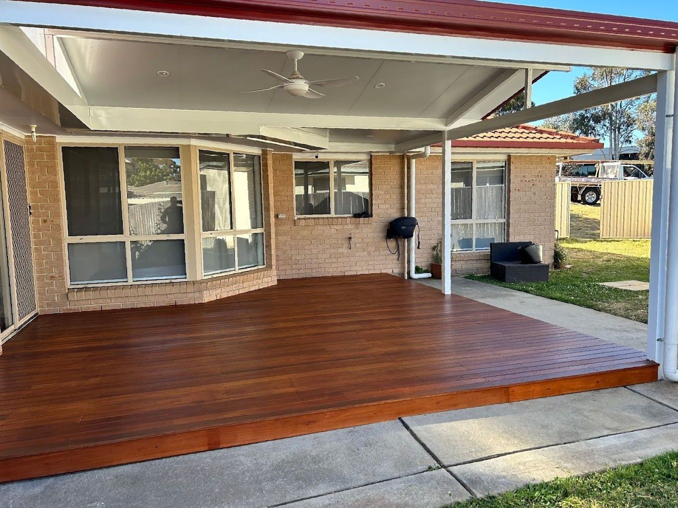 Covered wooden patio beside a brick house with white posts and sliding glass doors, viewed from the backyard
