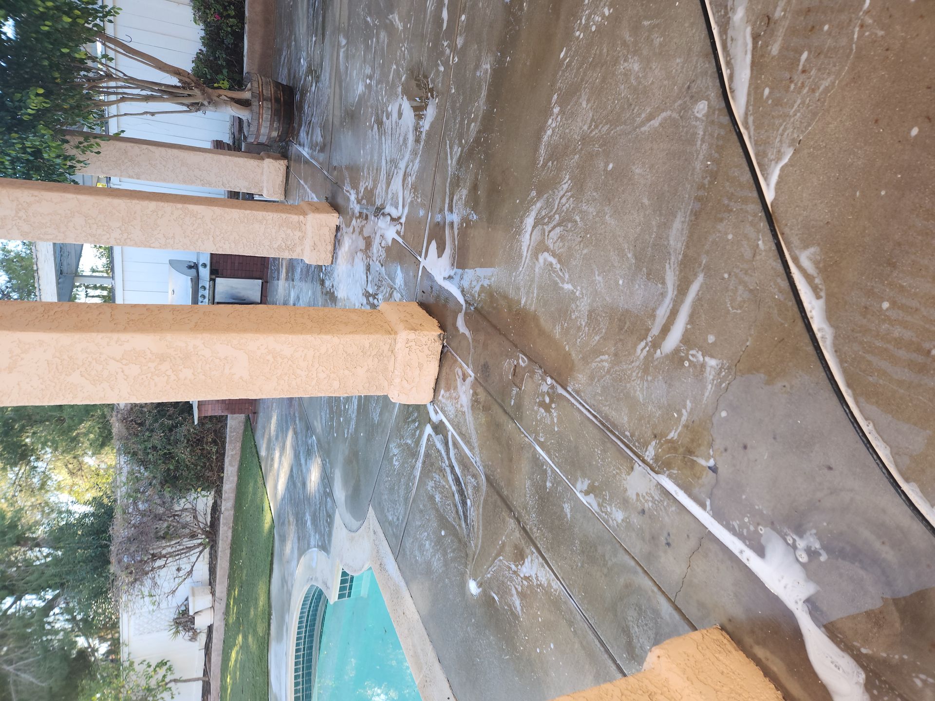 Wet concrete patio with tan columns, a pool edge, and trees in the background