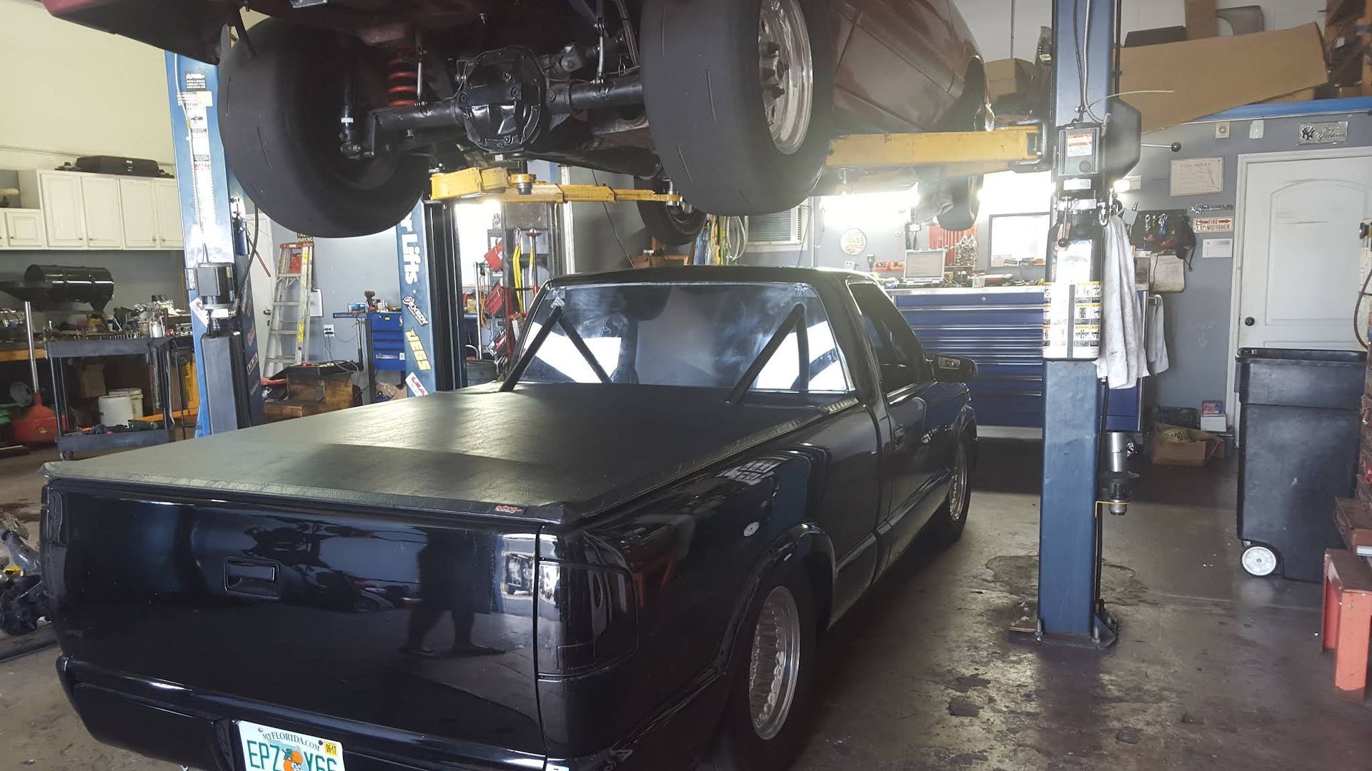 A black pickup truck parked inside a garage under a vehicle lifted by an automotive hydraulic rack.