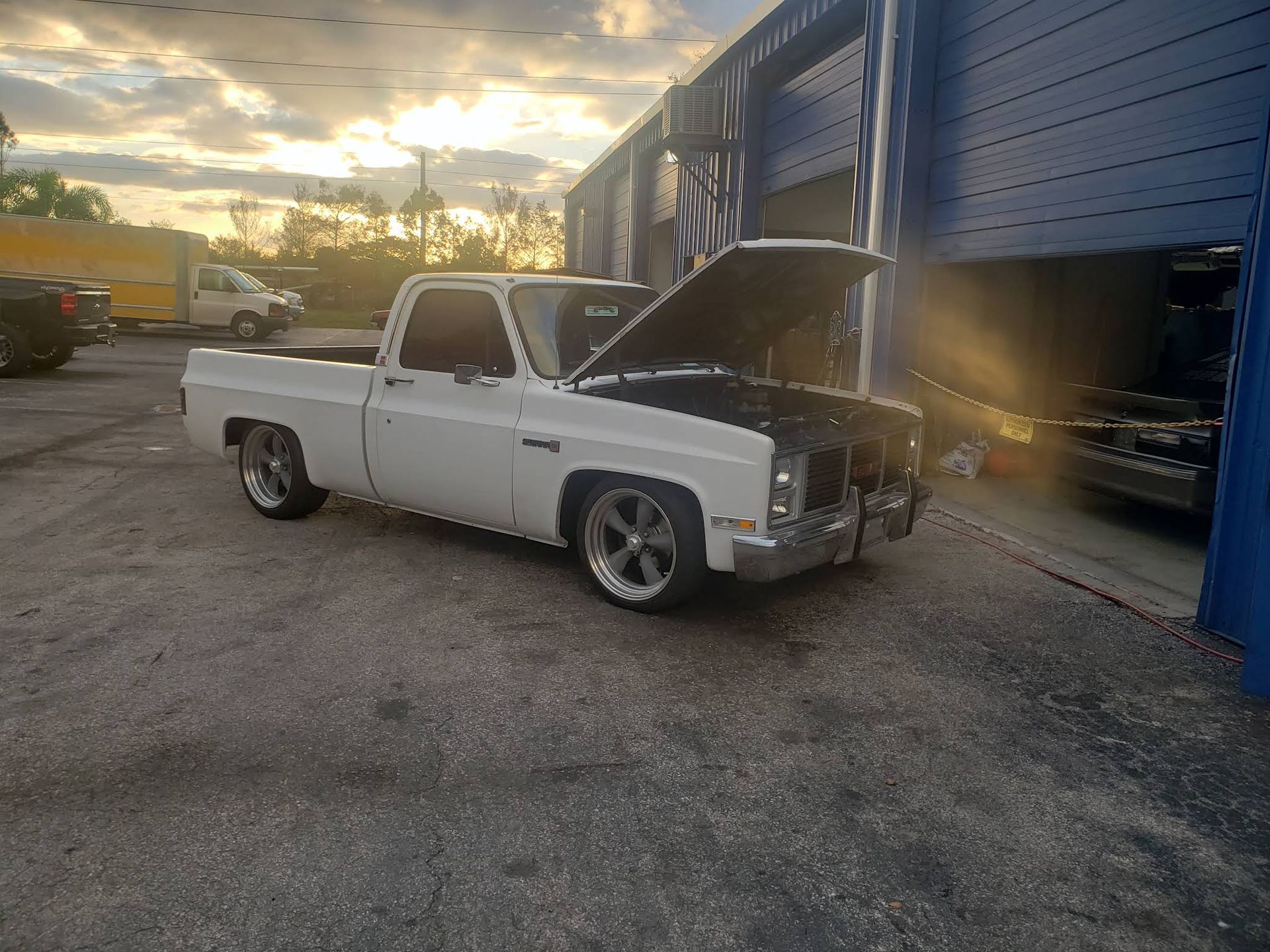 A white, lowered pickup truck with its hood open is parked on an asphalt lot in front of a blue industrial building.