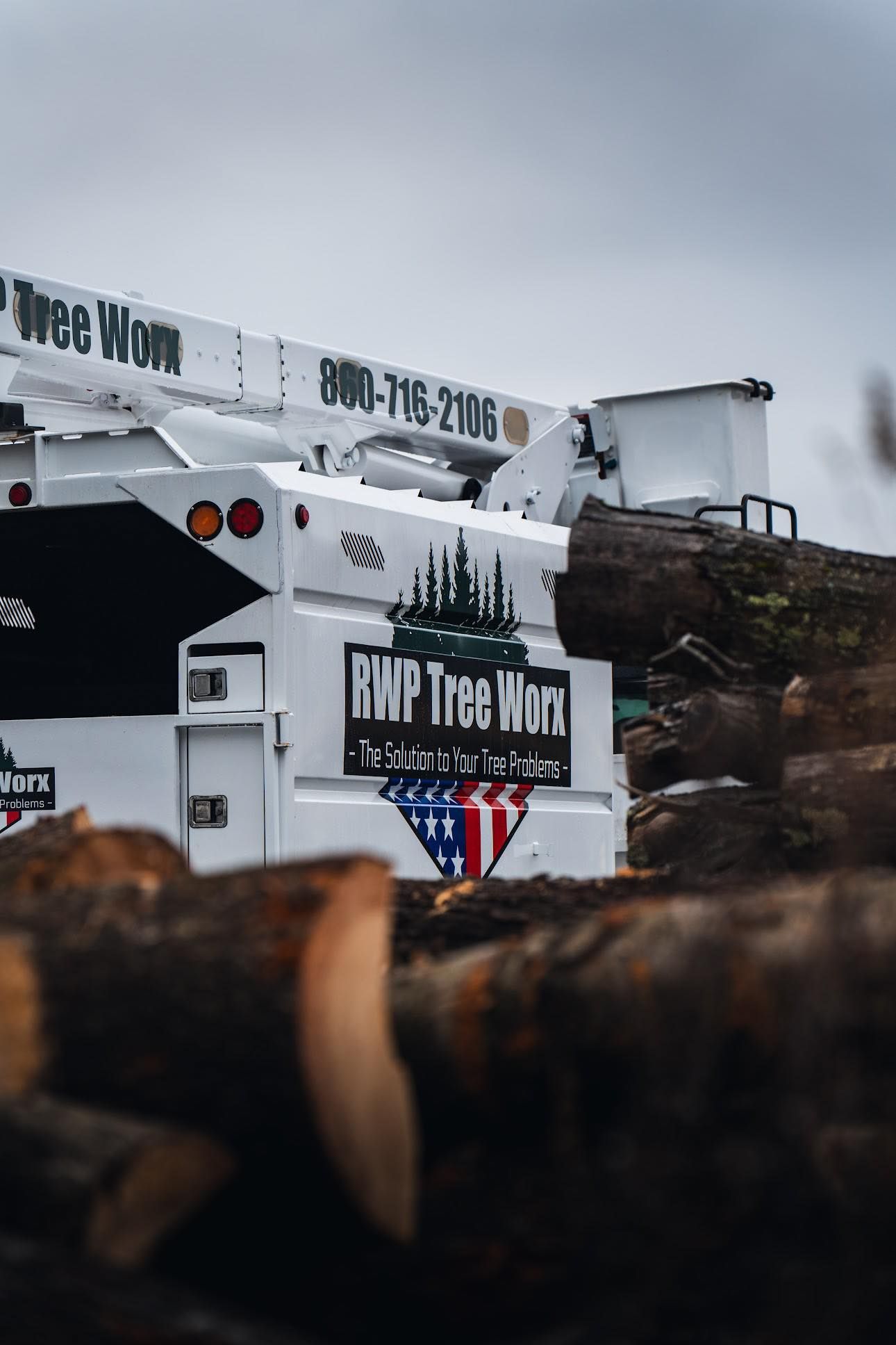 A white RWP Tree Work bucket truck parked near piles of logs under an overcast sky.