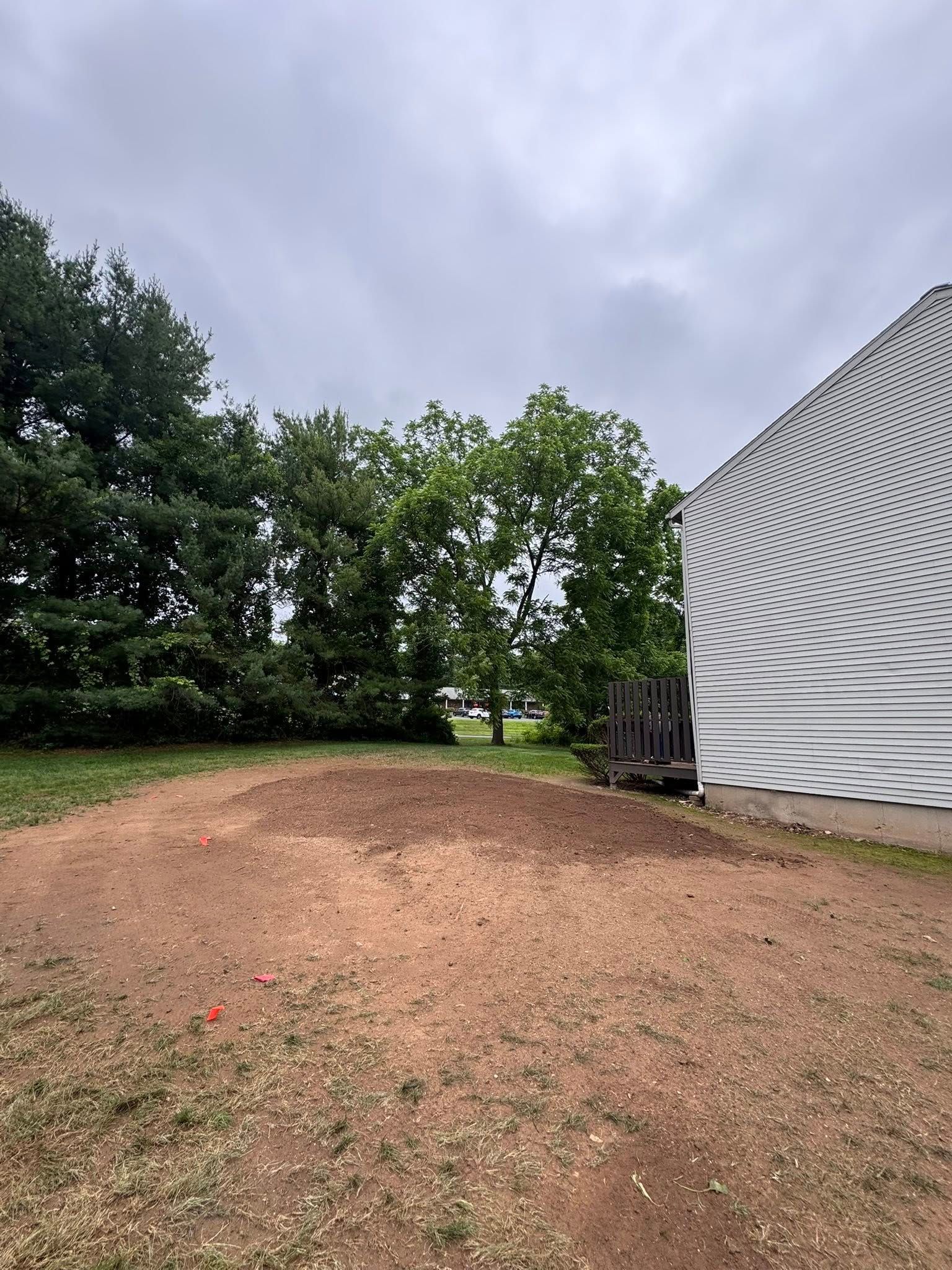 A pile of dirt sits in a yard next to a house with light-colored siding under a cloudy sky.