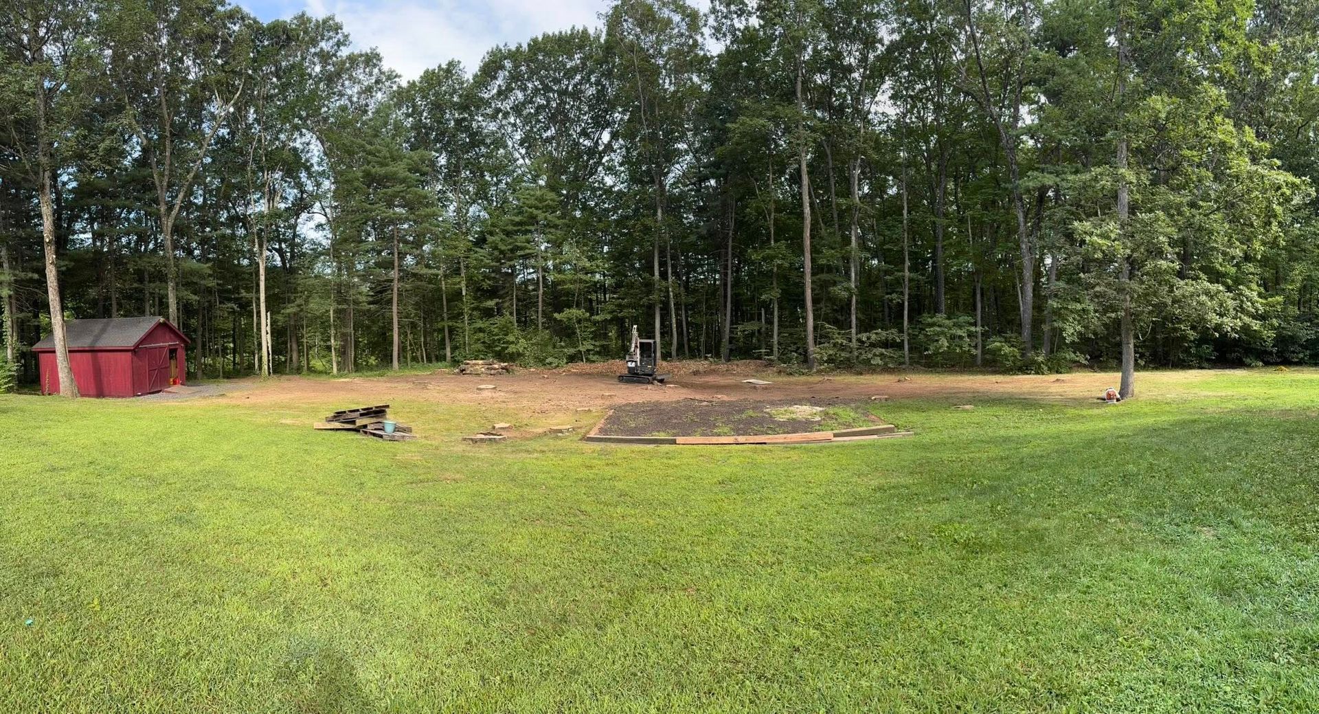 A small red shed sits on the edge of a cleared, dirt-filled construction area surrounded by a dense forest.