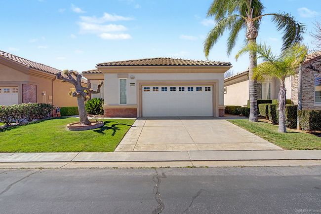 A one-story Spanish style house with two car garage. White garage, palm trees in the front and with a few clouds in blue sky.