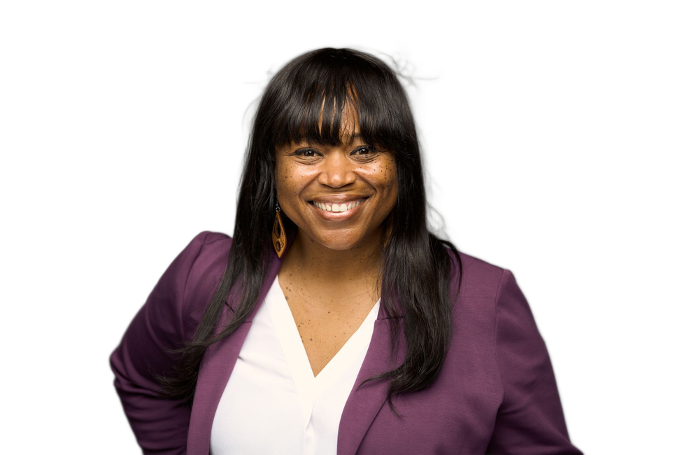Smiling person with hand on hip, wearing a purple blazer, white shirt, against a grey background.