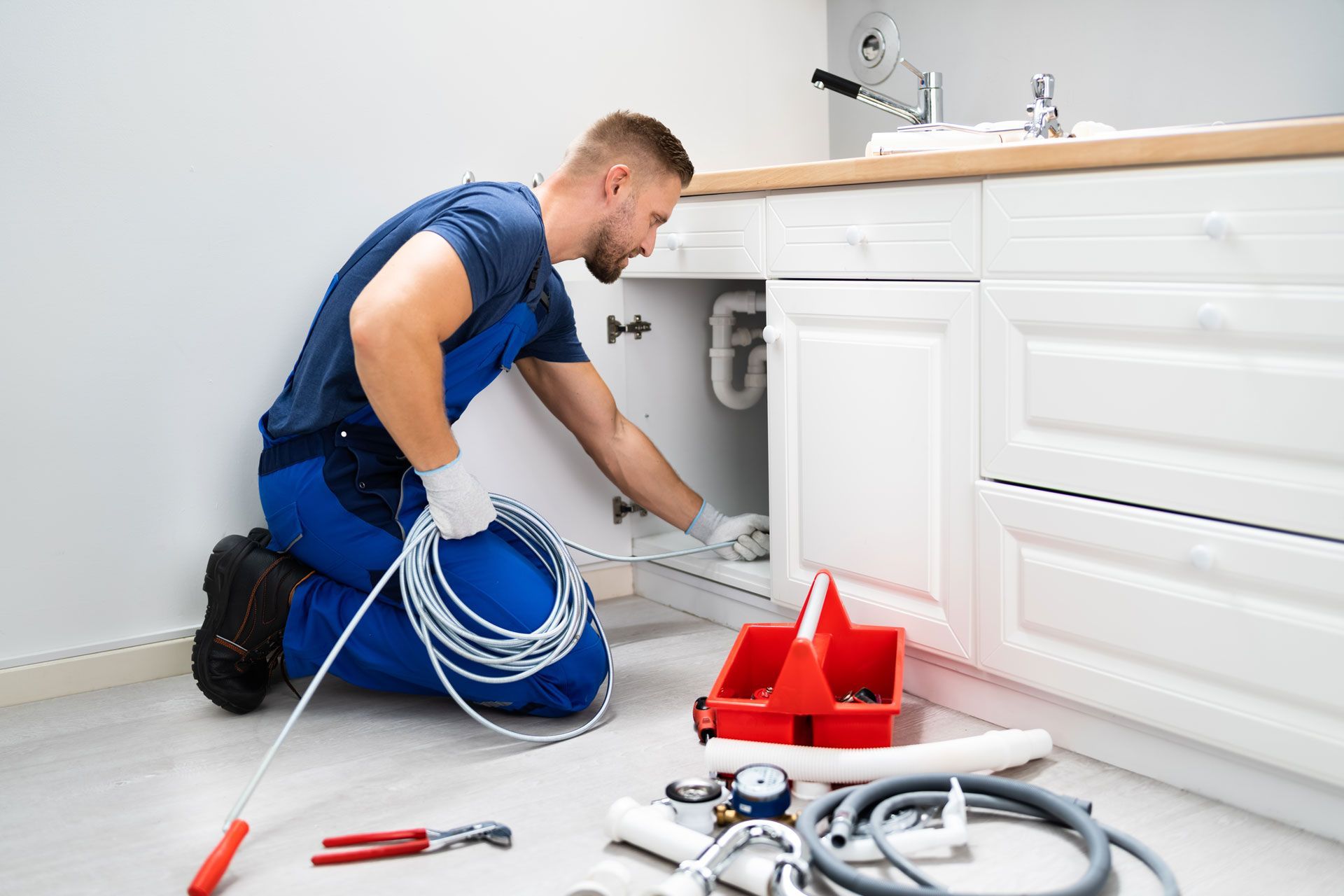 Plumber in blue coveralls kneeling under a kitchen sink, using a drain snake.