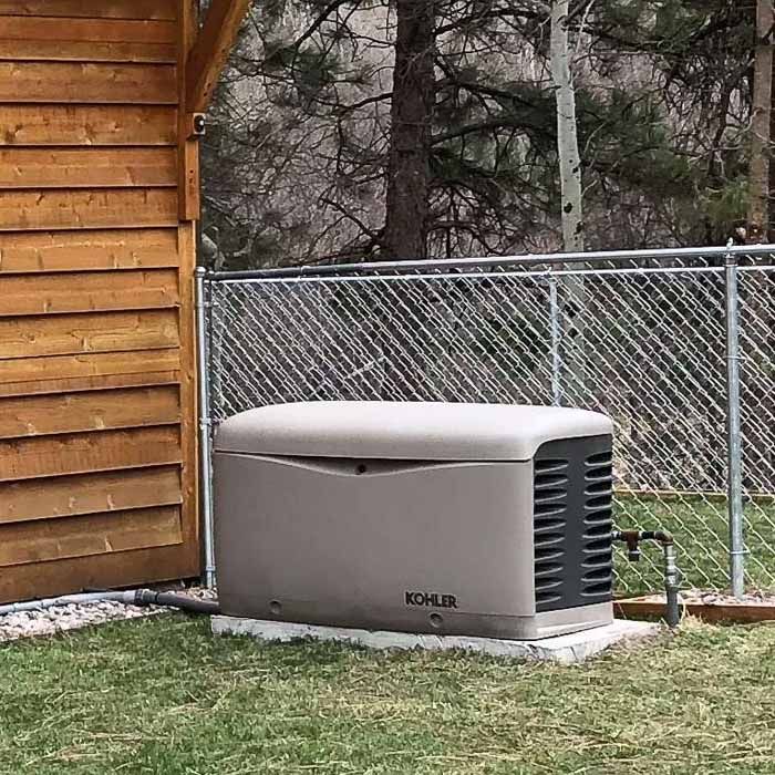 A Kohler generator, beige casing, sits outside, near a wooden structure and chain-link fence.