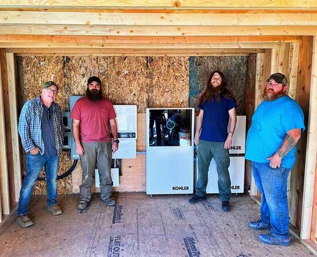 Four people stand inside a wooden utility shed next to an installed Kohler home generator and electrical equipment.