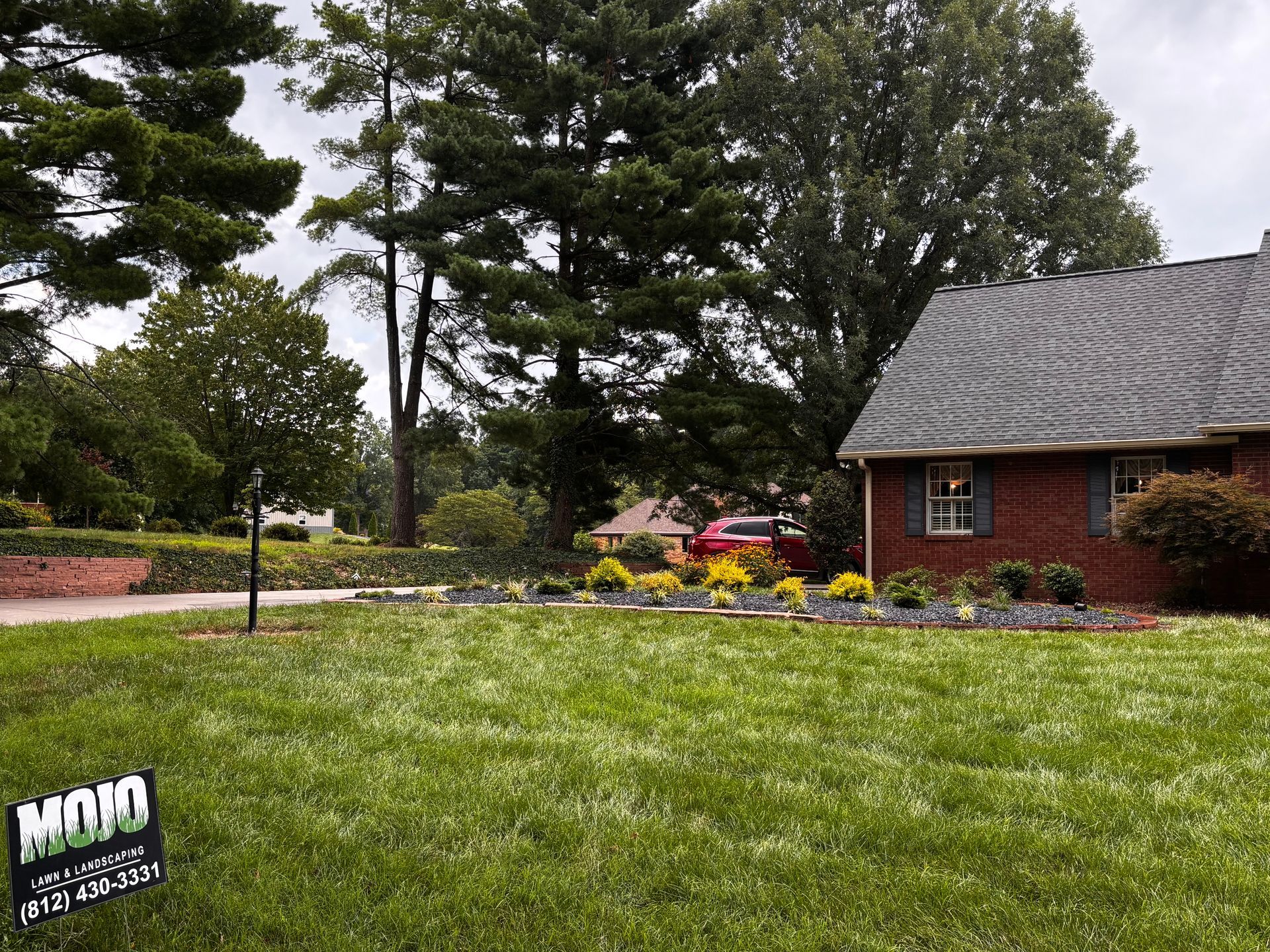 Suburban yard with green lawn, trees, flower bed, and a brick house under cloudy sky
