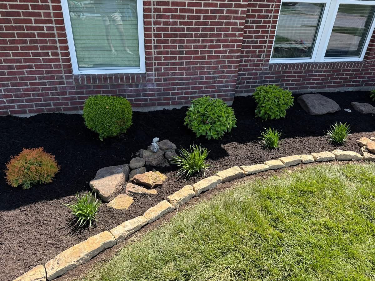 Curved brick flower bed with small shrubs and rocks beside a lawn and brick house wall