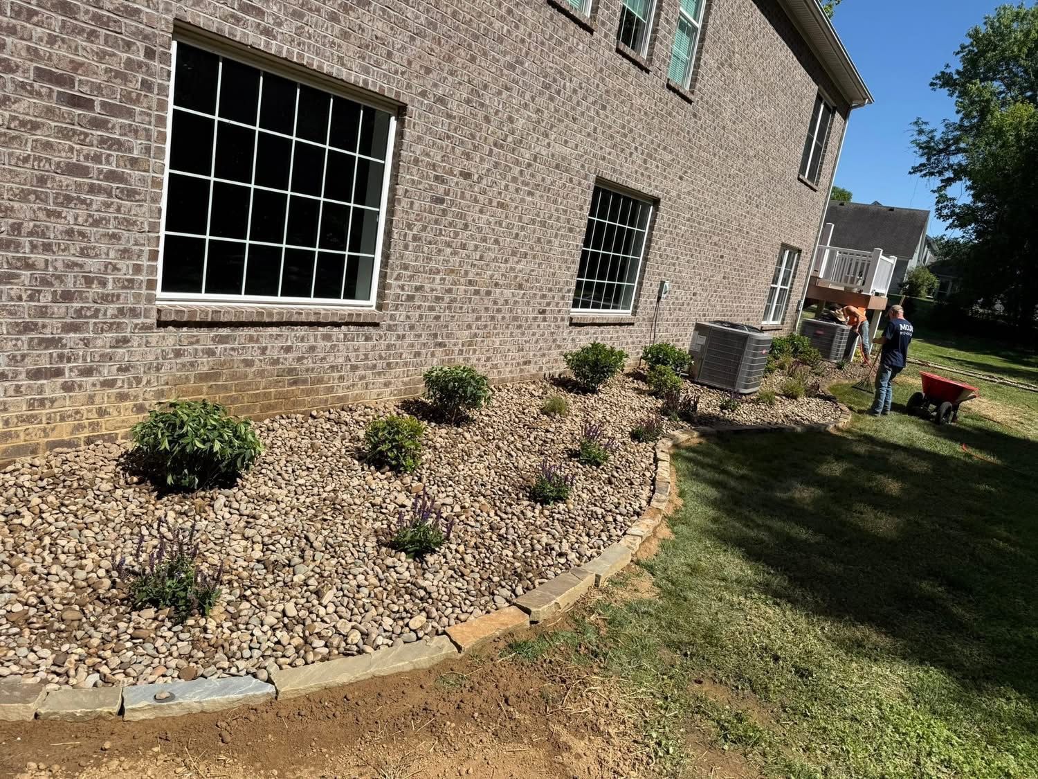 Brick house side yard with new mulch and small shrubs, bordered by grass and a patio chair near the corner