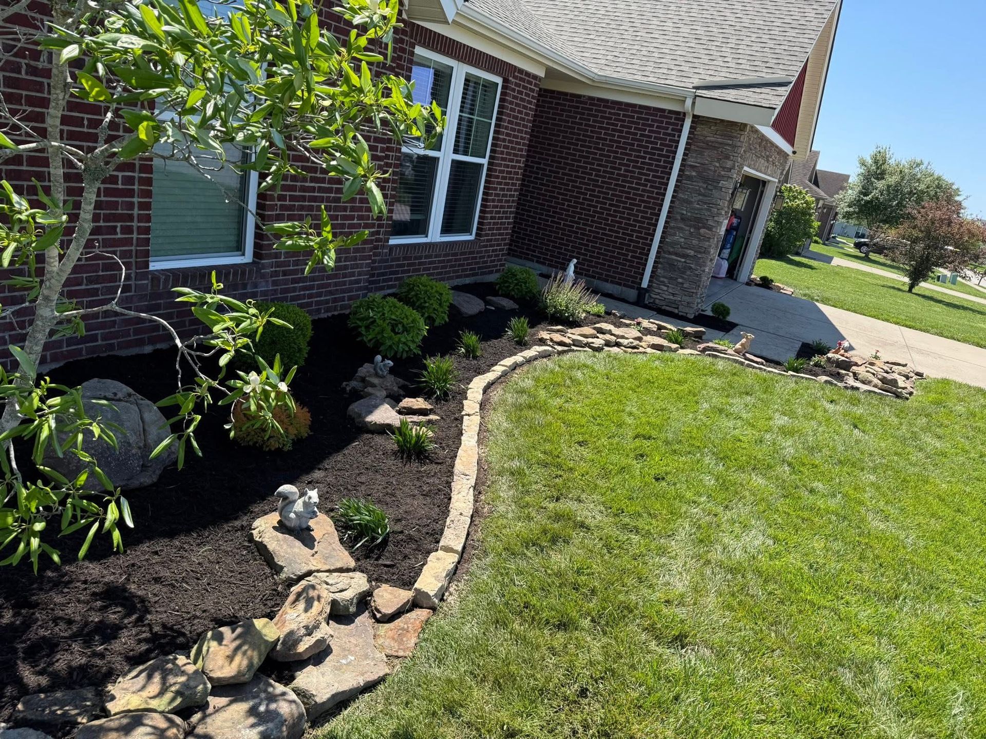 Curved stone garden bed beside a brick house, with black mulch and green shrubs along a lawn.