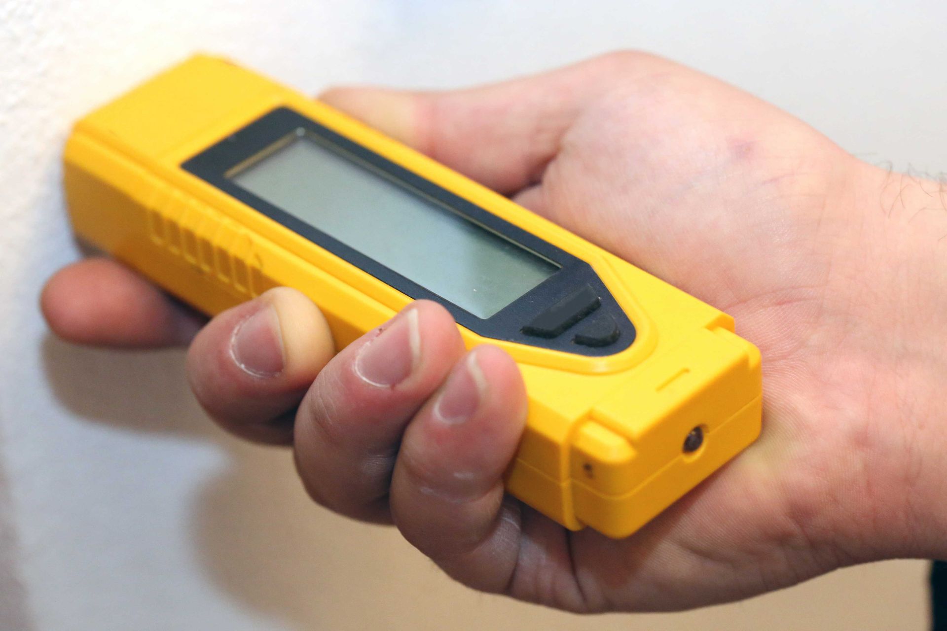 Hand holding a yellow stud finder against a white wall.