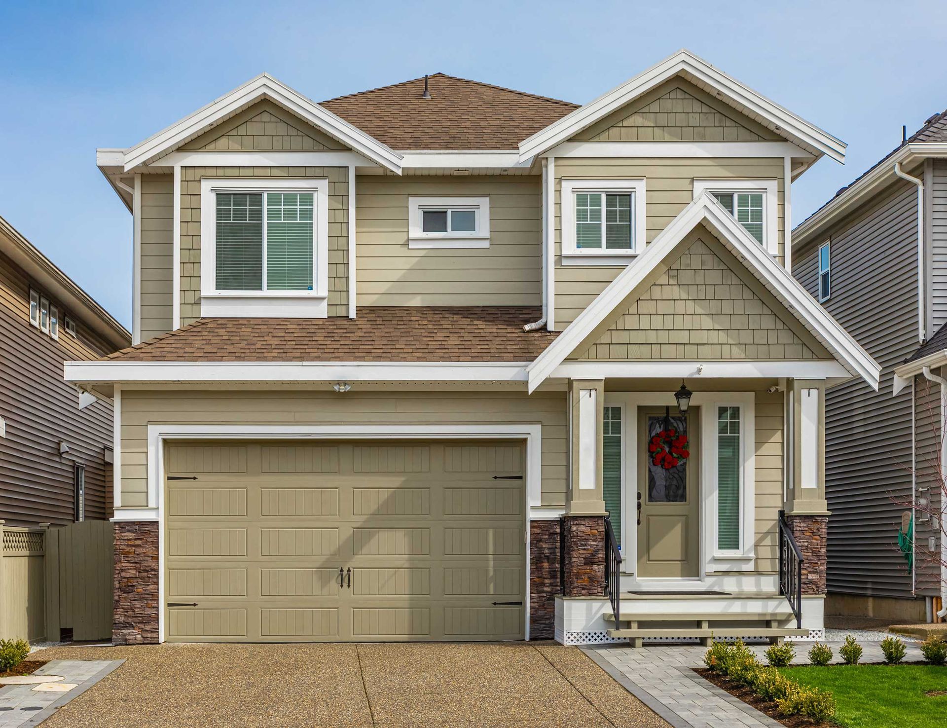 Two-story house with tan siding, brown roof, and attached garage. Front door has a red wreath.