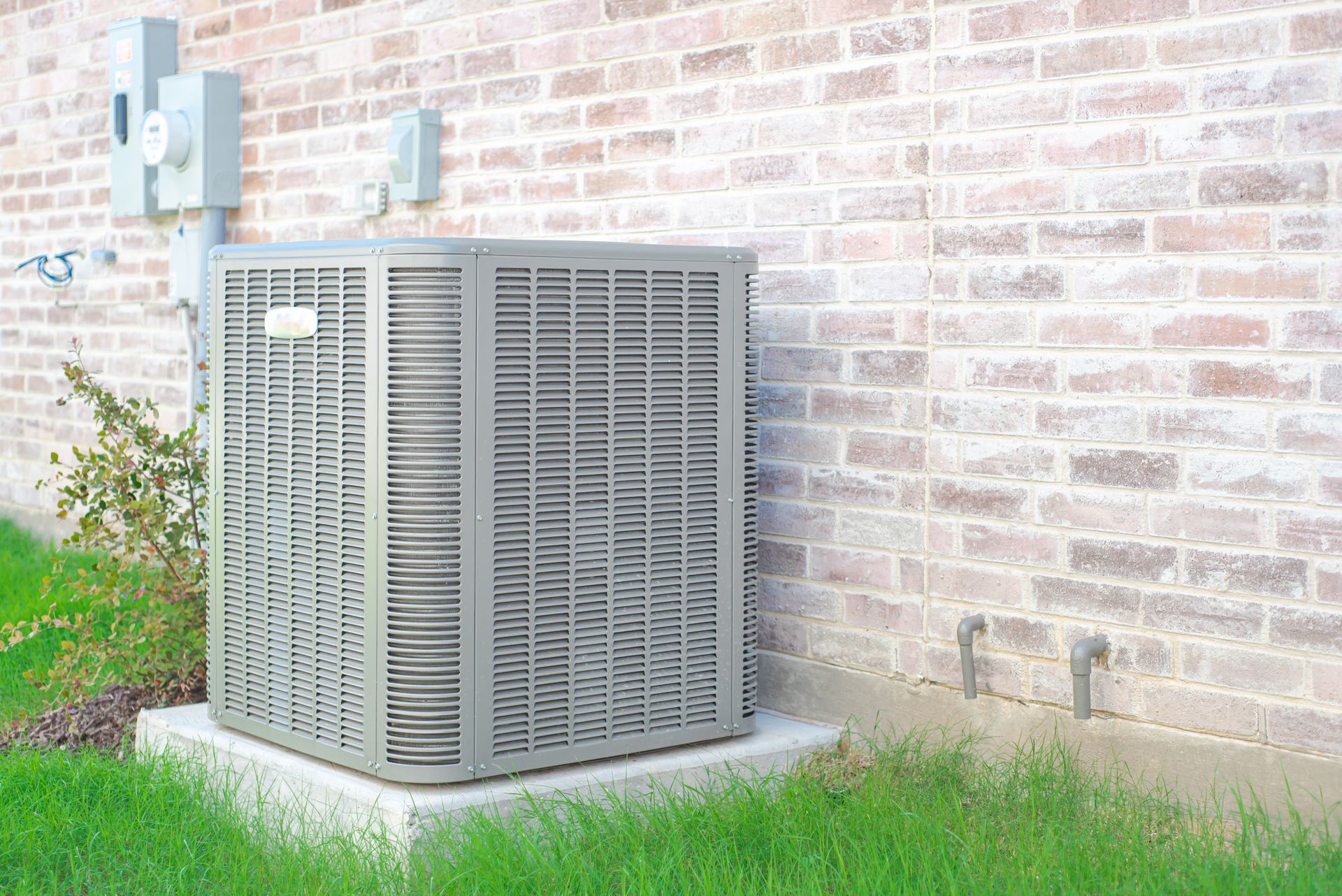 Outdoor HVAC unit beside a brick wall, sitting on a concrete pad near grass and shrubs