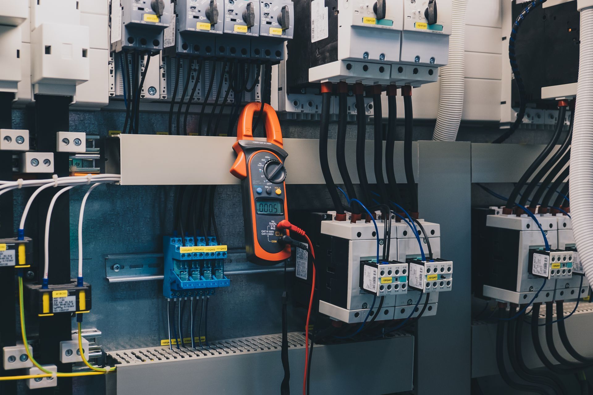 Industrial electrical control panel with wires, breakers, and a handheld multimeter mounted inside a cabinet