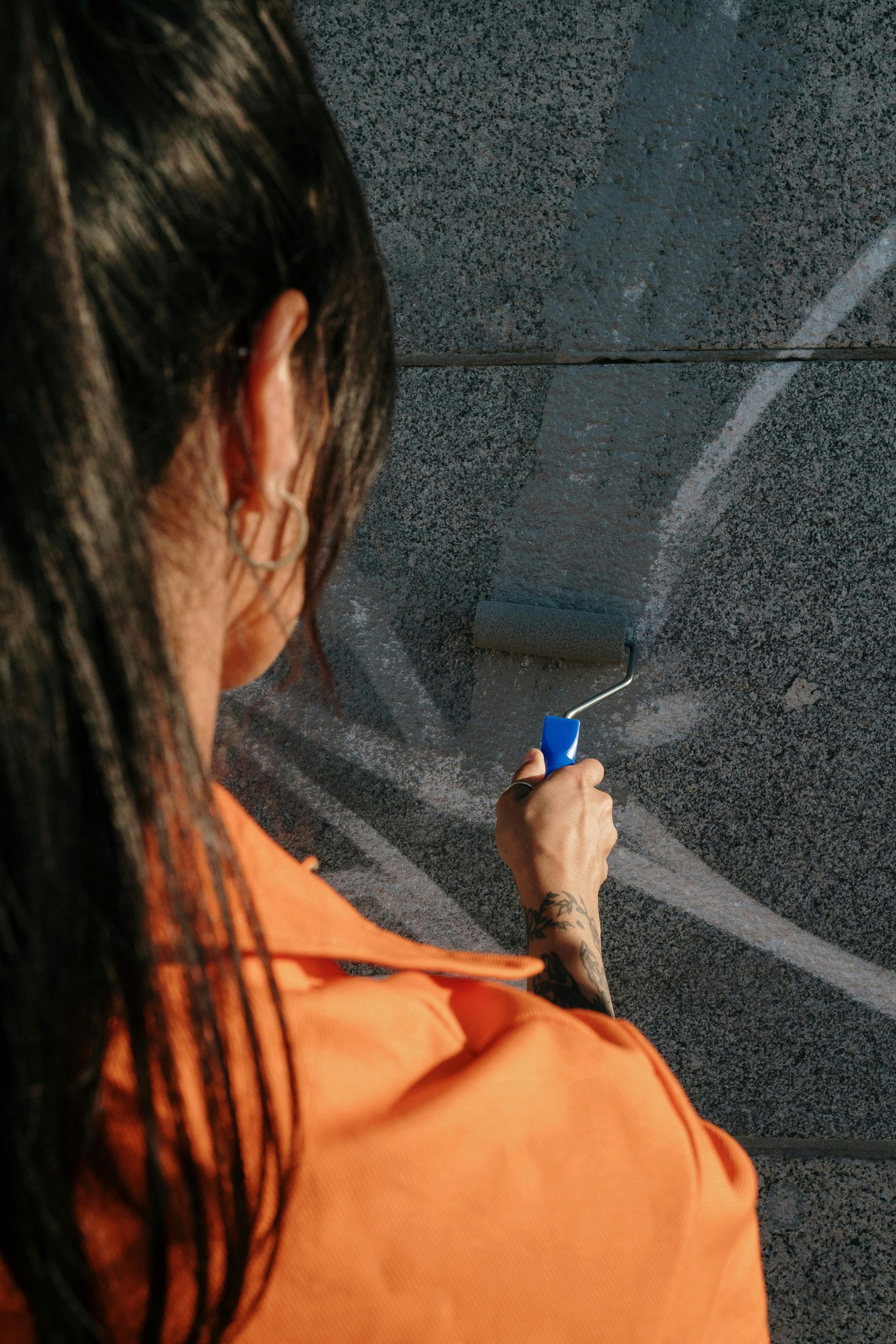 Person in orange jumpsuit using a paint roller to cover white graffiti on a textured gray wall.